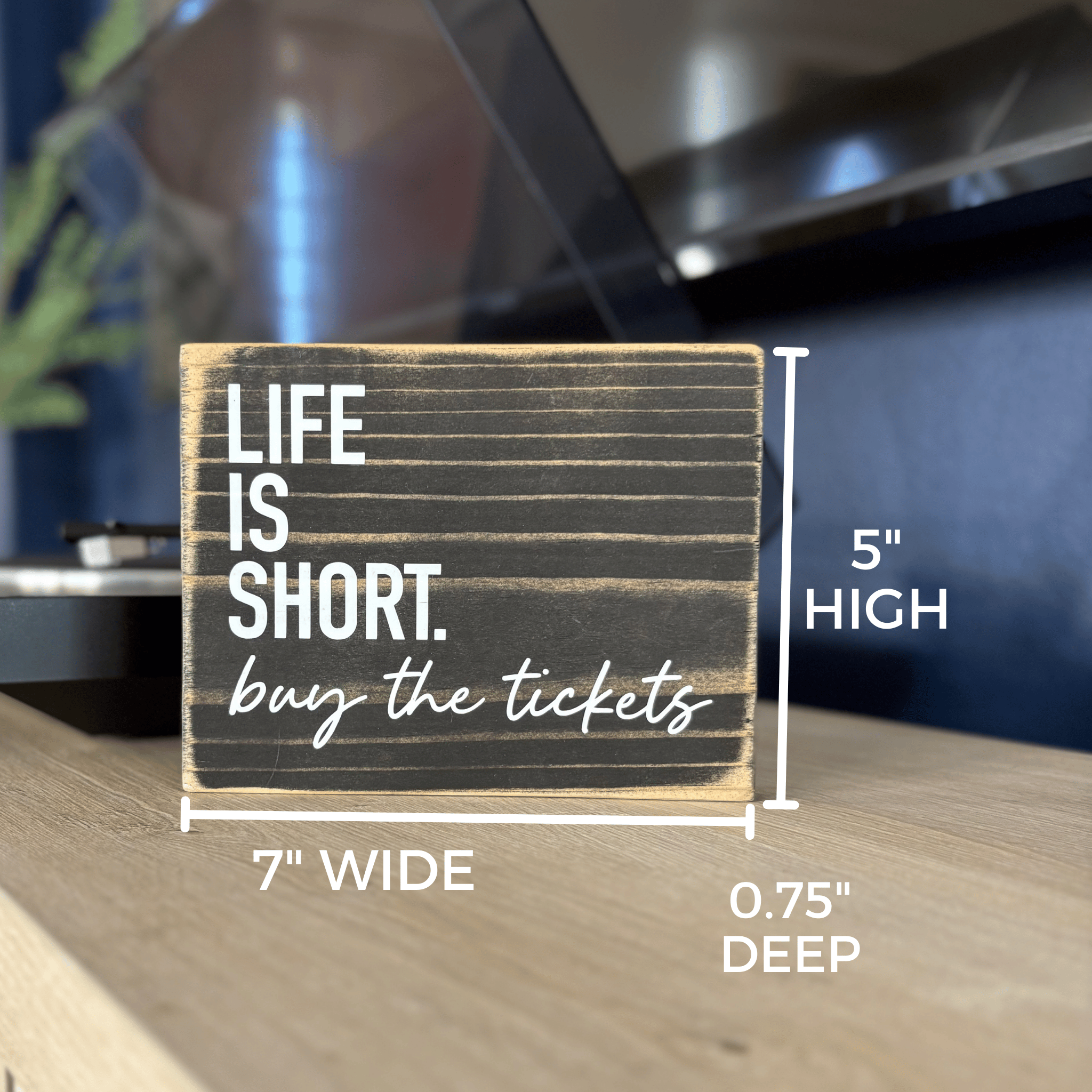 a black sign sits on a table next to a record player, it says Life is Short. Buy the tickets. Measurements indicate its size.