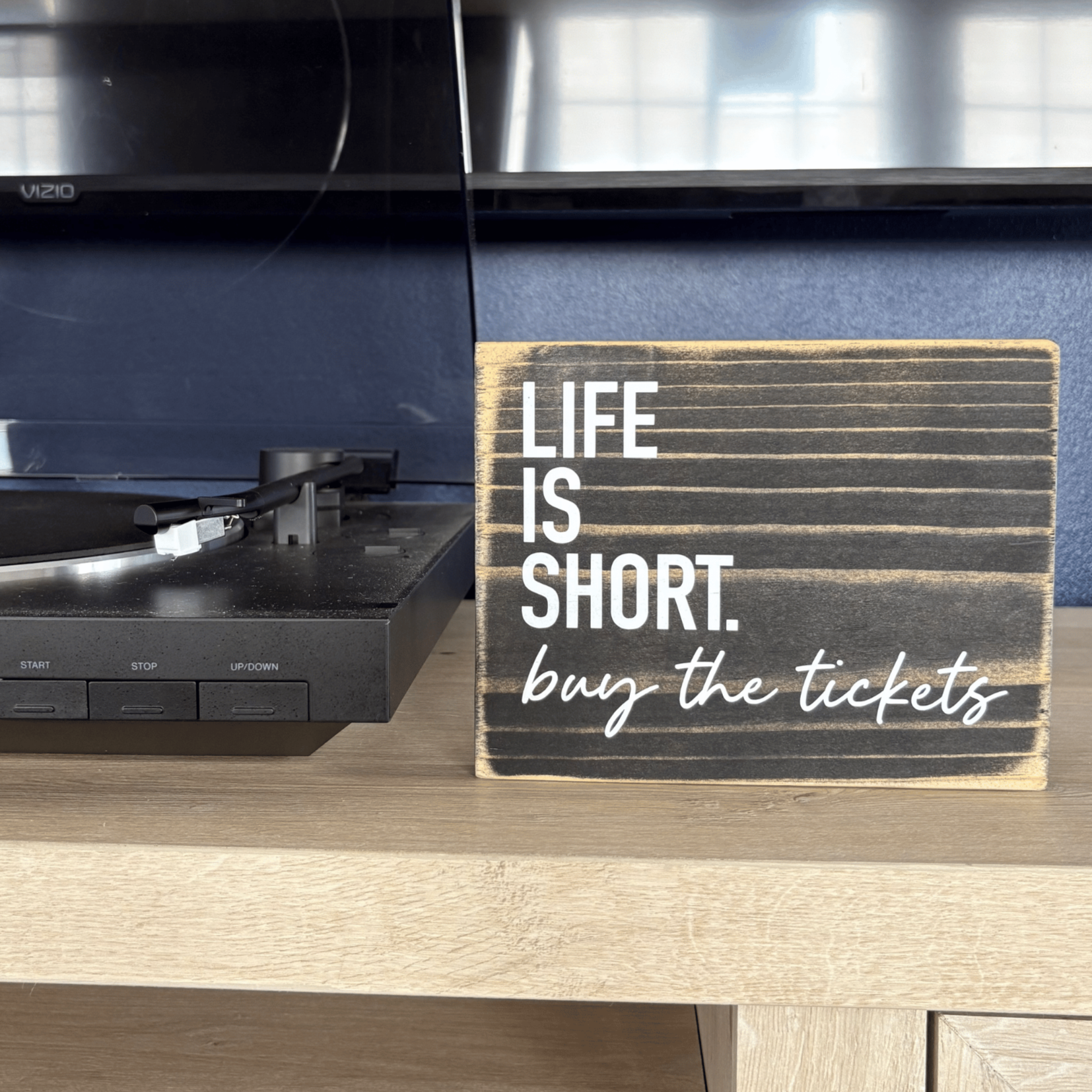 a black sign sits on a table next to a record player, it says Life is Short. Buy the tickets.