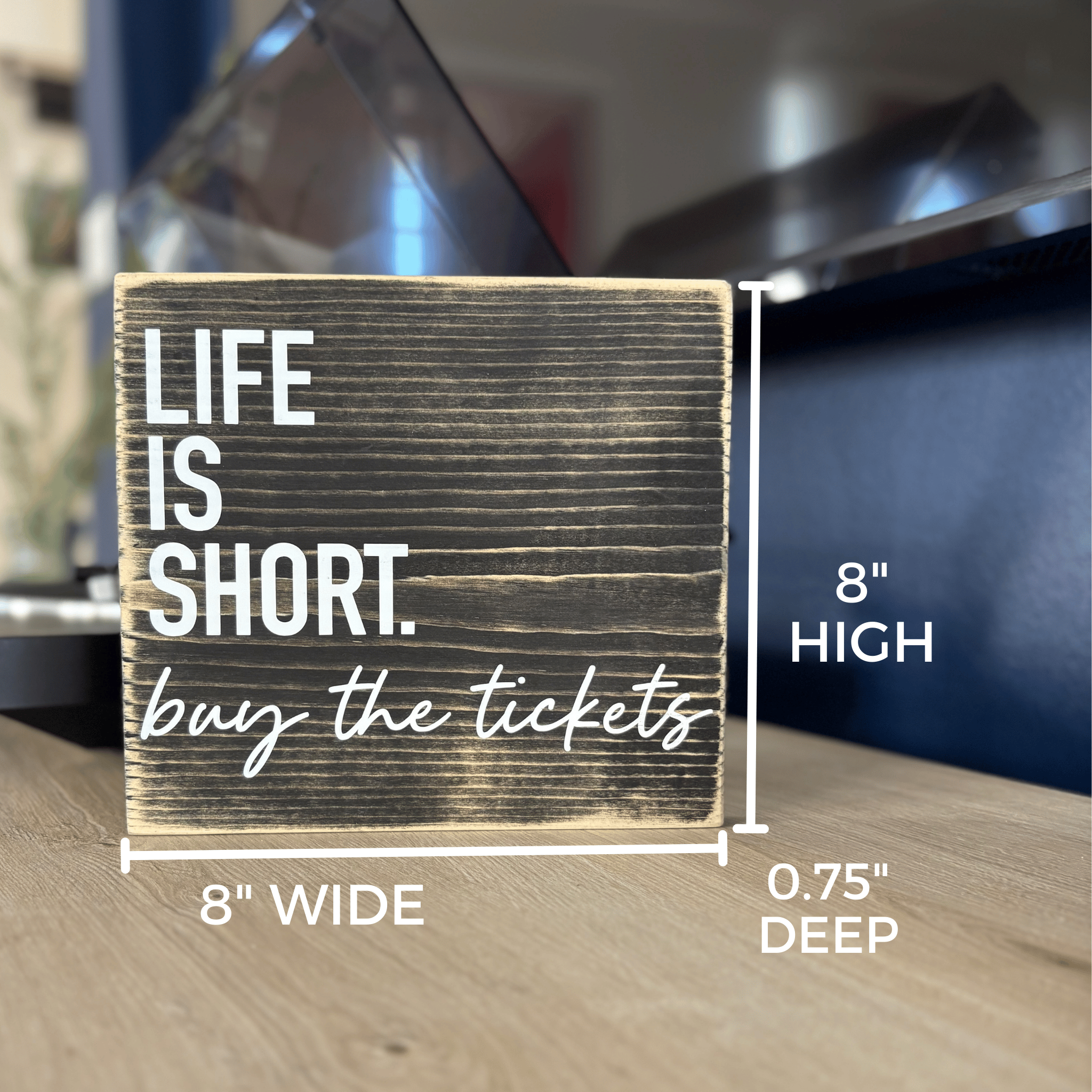 Wooden sign with 'Life is short, buy the tickets' text and dimensions on a blurred background