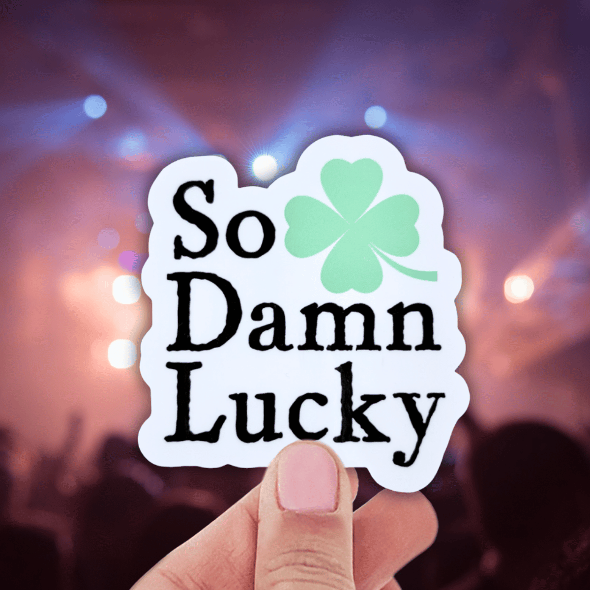 Sticker with 'So Damn Lucky' text and a clover leaf held by a hand against a blurred background.