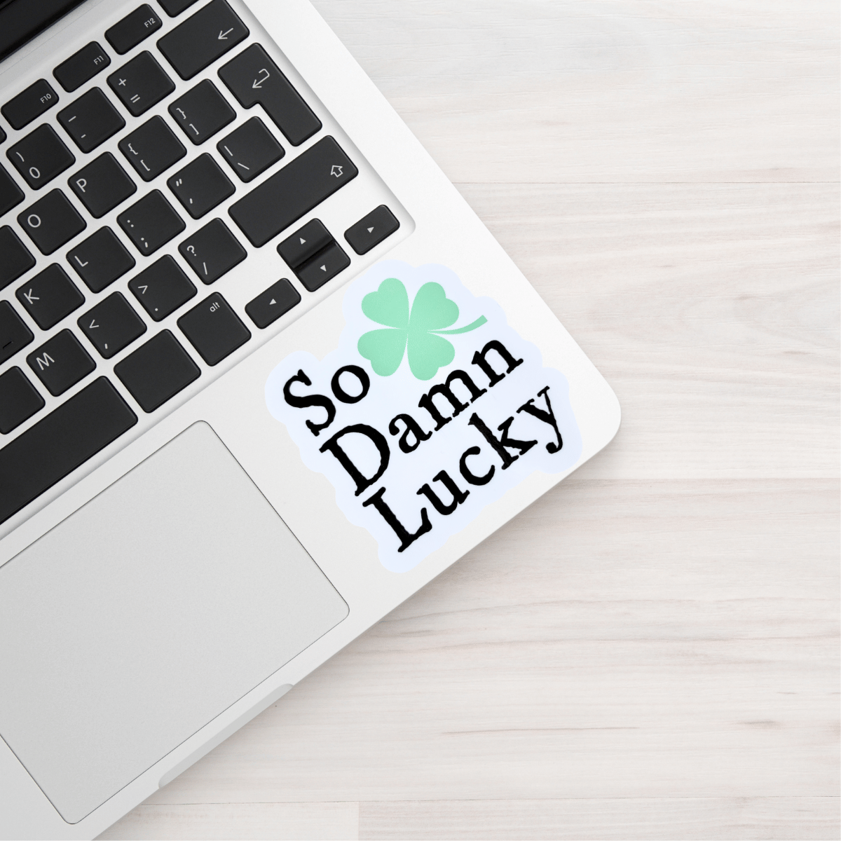 Laptop with a sticker reading 'So Damn Lucky' featuring a four-leaf clover on a light wooden surface.