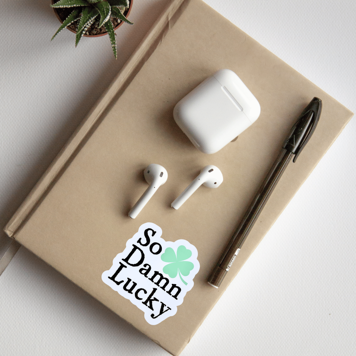 White AirPods, white case, pen, and 'So Damn Lucky' sticker on a beige notebook with a plant in the corner.