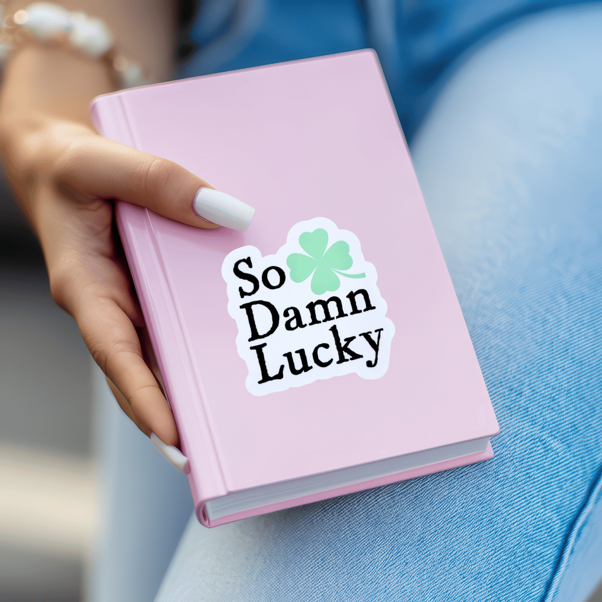Pink notebook with 'So Damn Lucky' sticker held by a person wearing blue jeans.