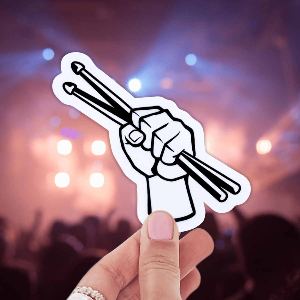 Hand holding a sticker of a hand with drumsticks against a concert background