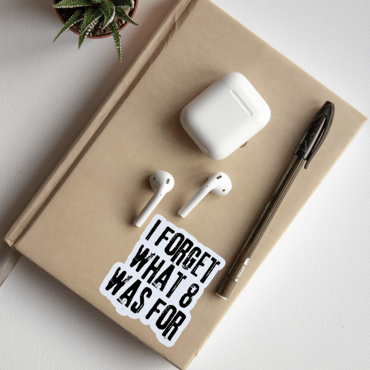 AirPods, white case, pen, and 'I FORGET WHAT 8 WAS FOR' sticker on a beige notebook.
