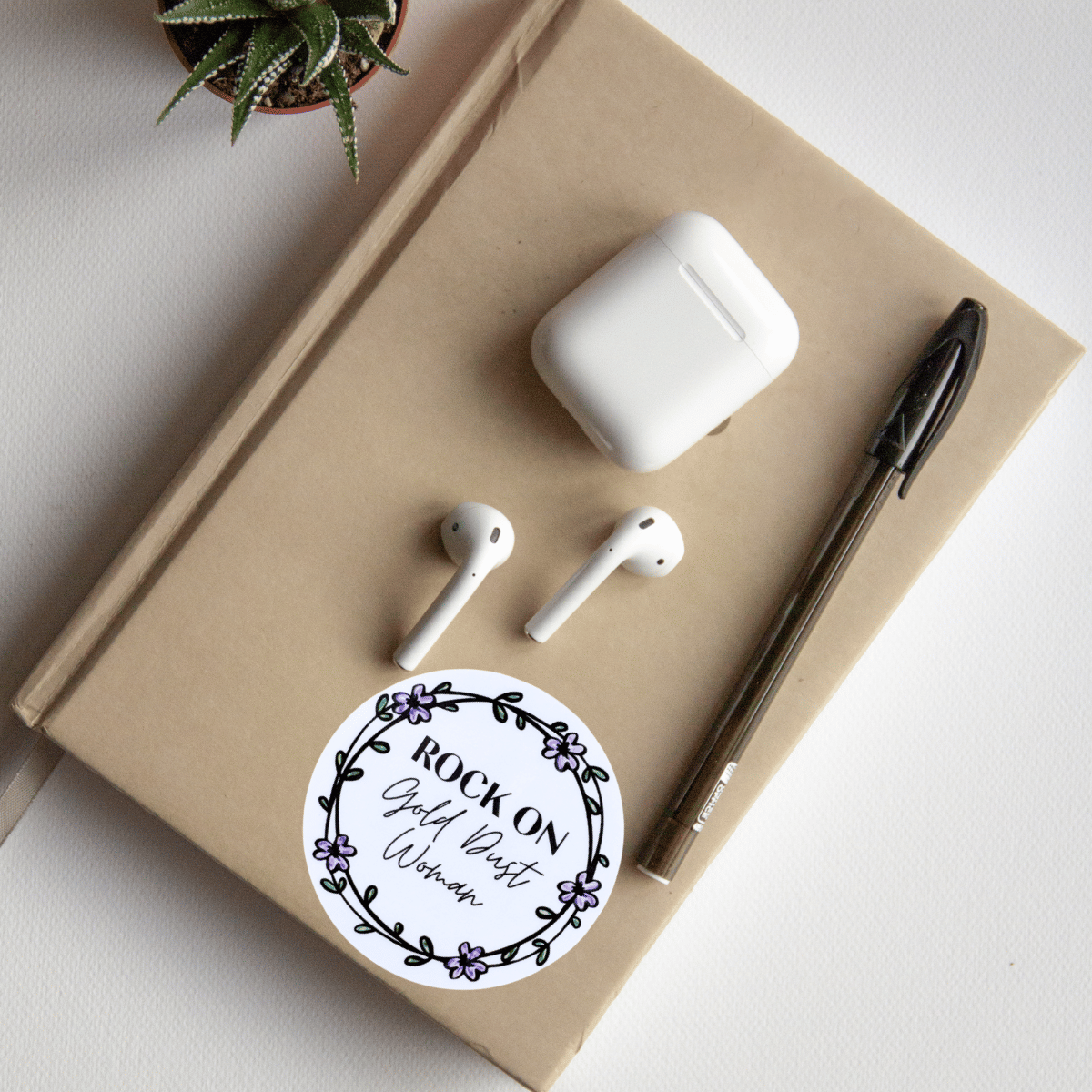 White wireless earbuds and case on a beige notebook with a sticker and pen, on a light gray surface.