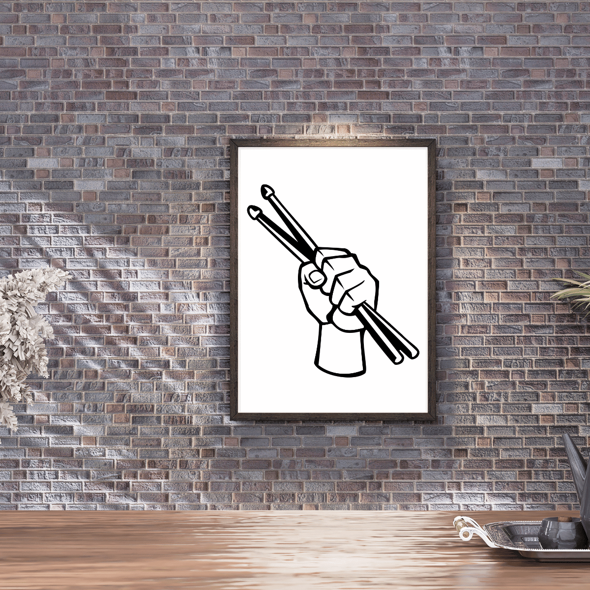 A framed black and white graphic art print of a hand holding drumsticks, displayed on a rustic gray brick wall above a wooden table.