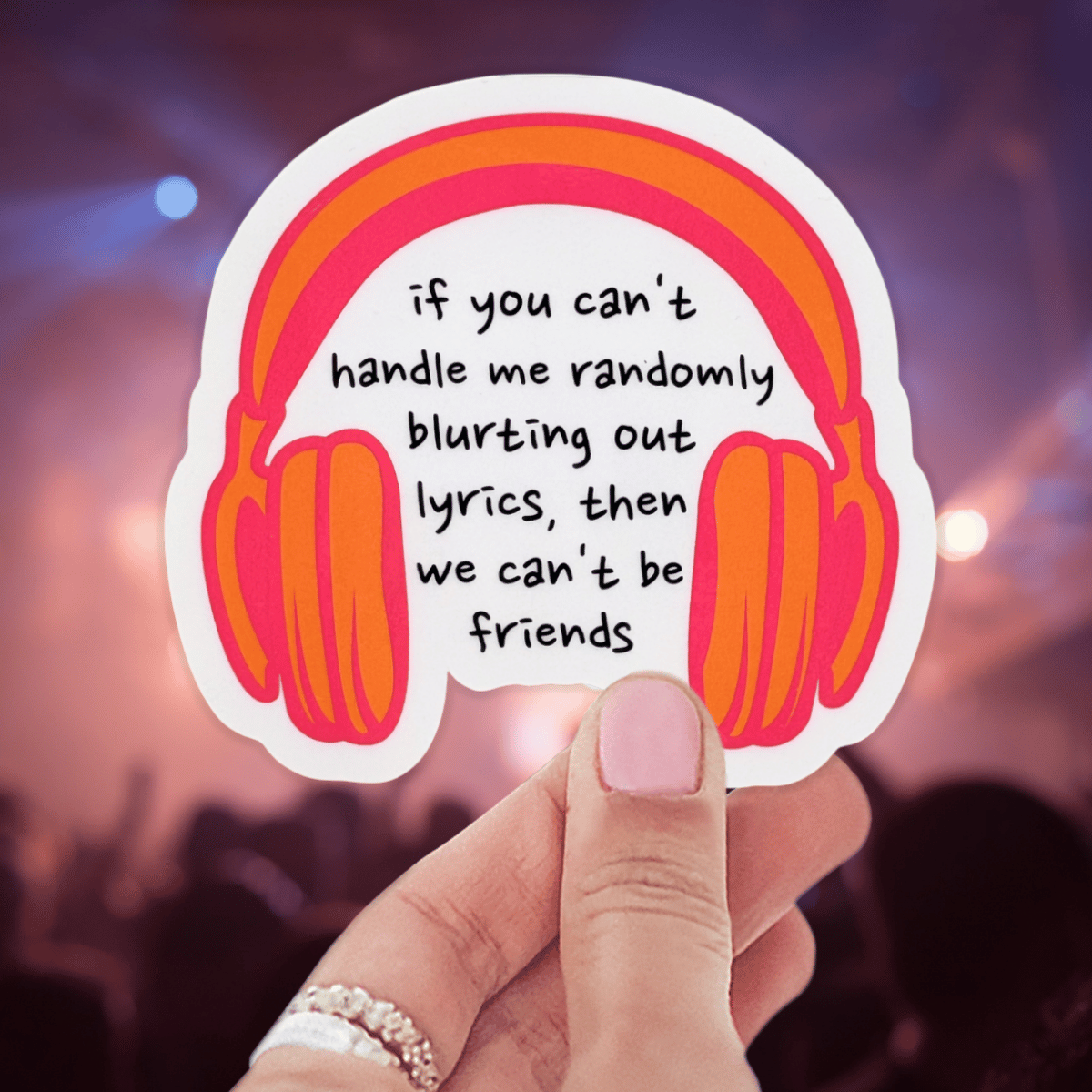 Hand holding a sticker with headphones and text against a blurred concert background