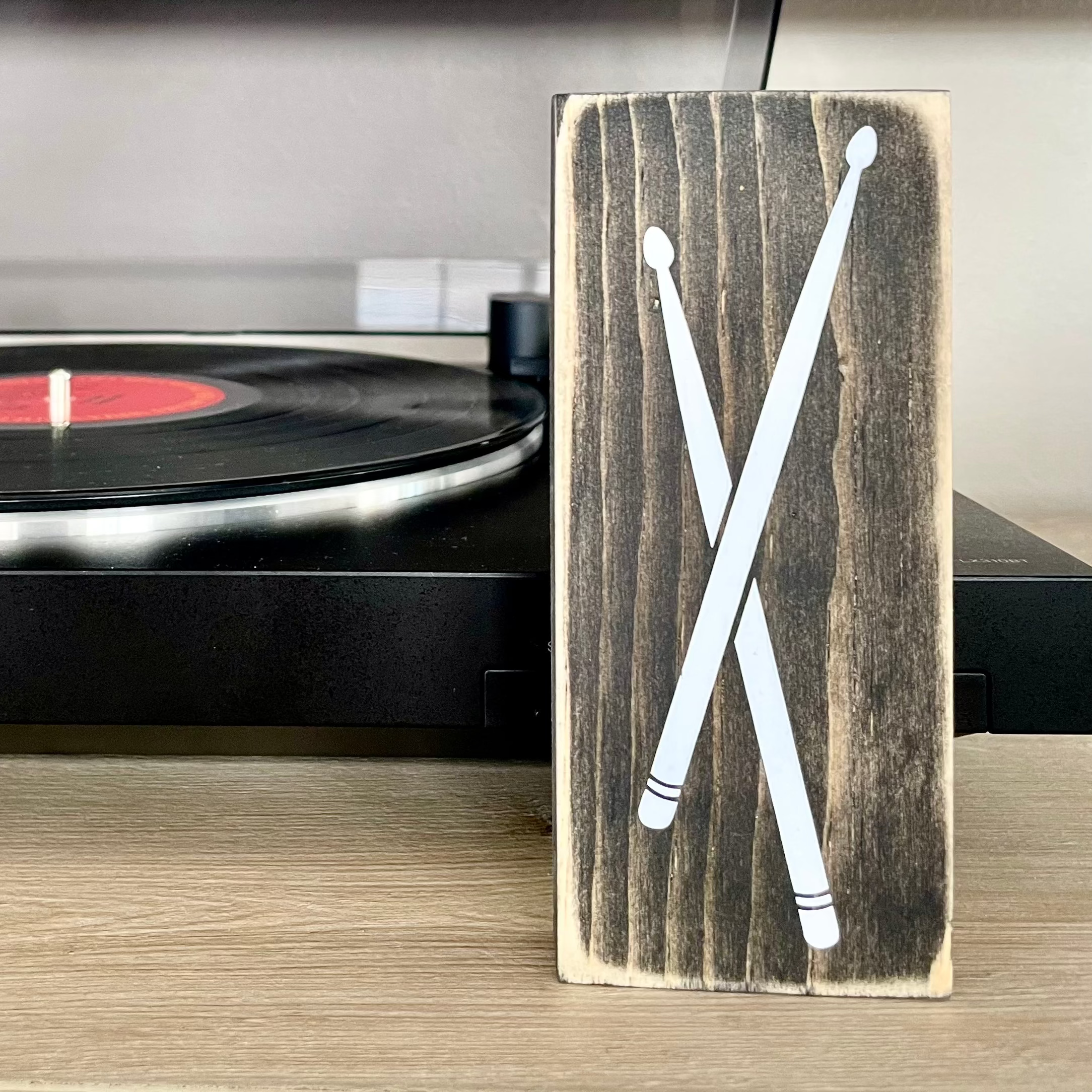 A small, black, wood sign with drumsticks painted on it, sits vertically on a table in front of a record player.