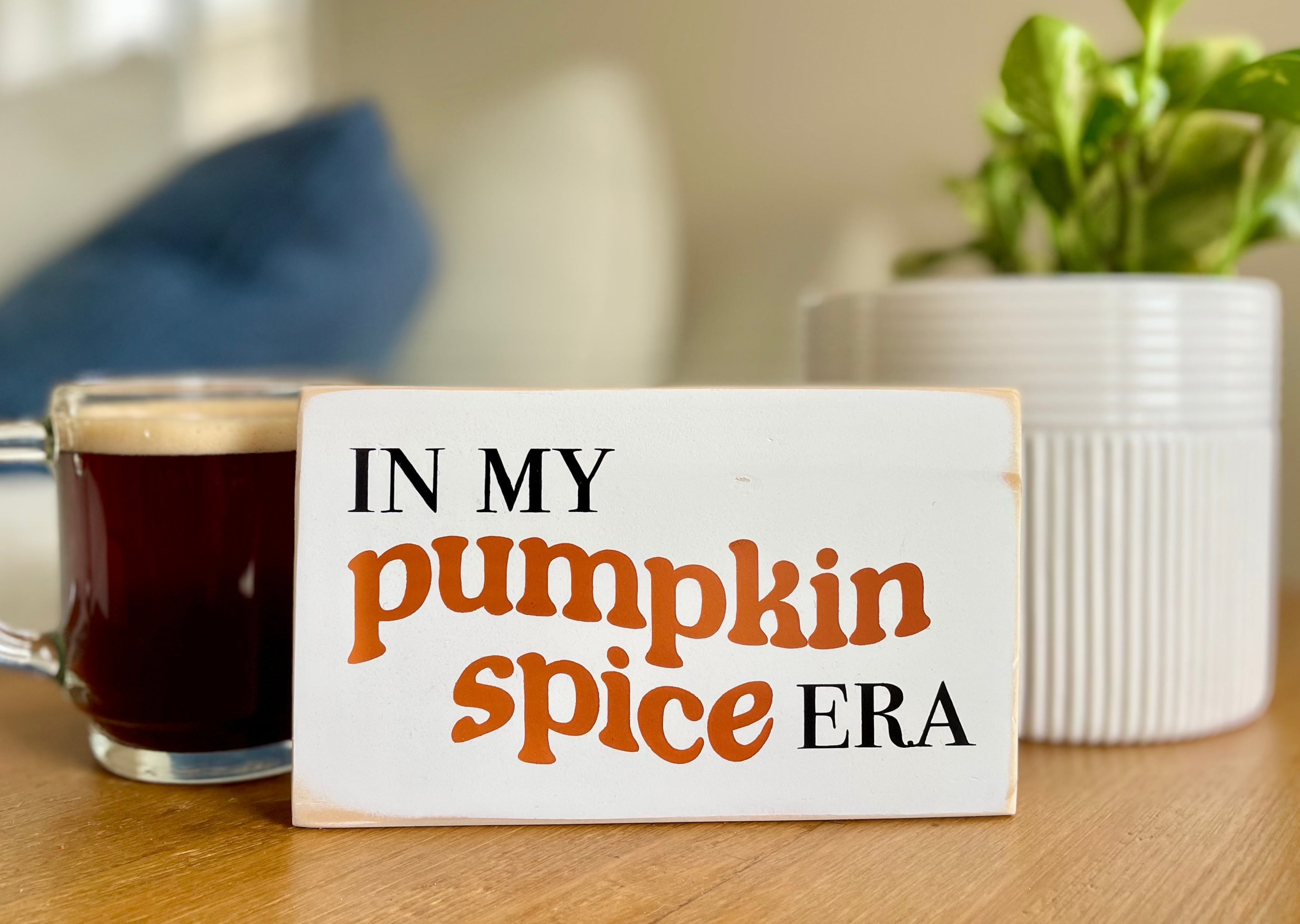 a small, white, wood sign that reads" in my pumpkin spice era" sits on a table with a cup of coffee and a plant next to it.