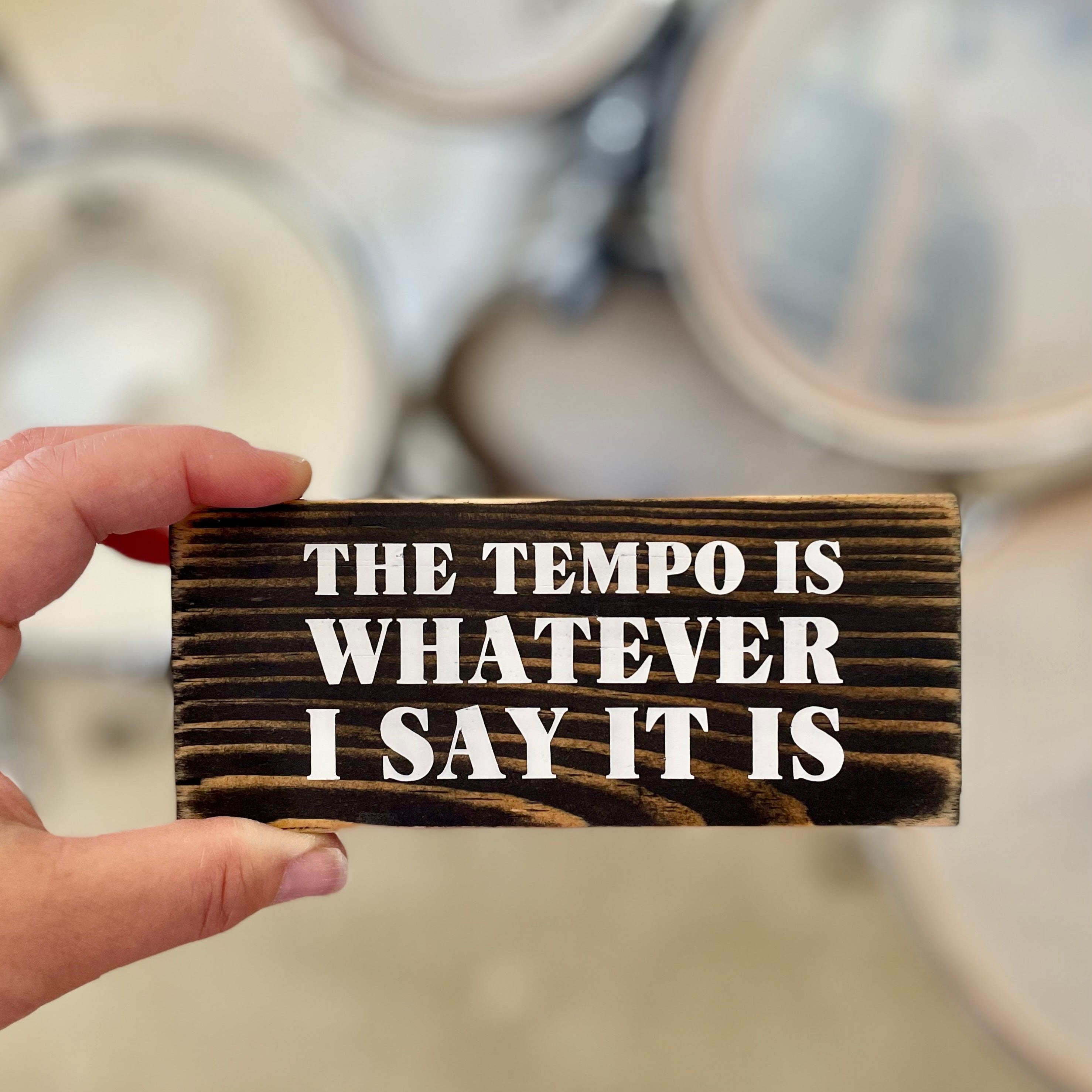 A small, black, wood sign is being held in someone's hand. A drum kit is blurry in the background.