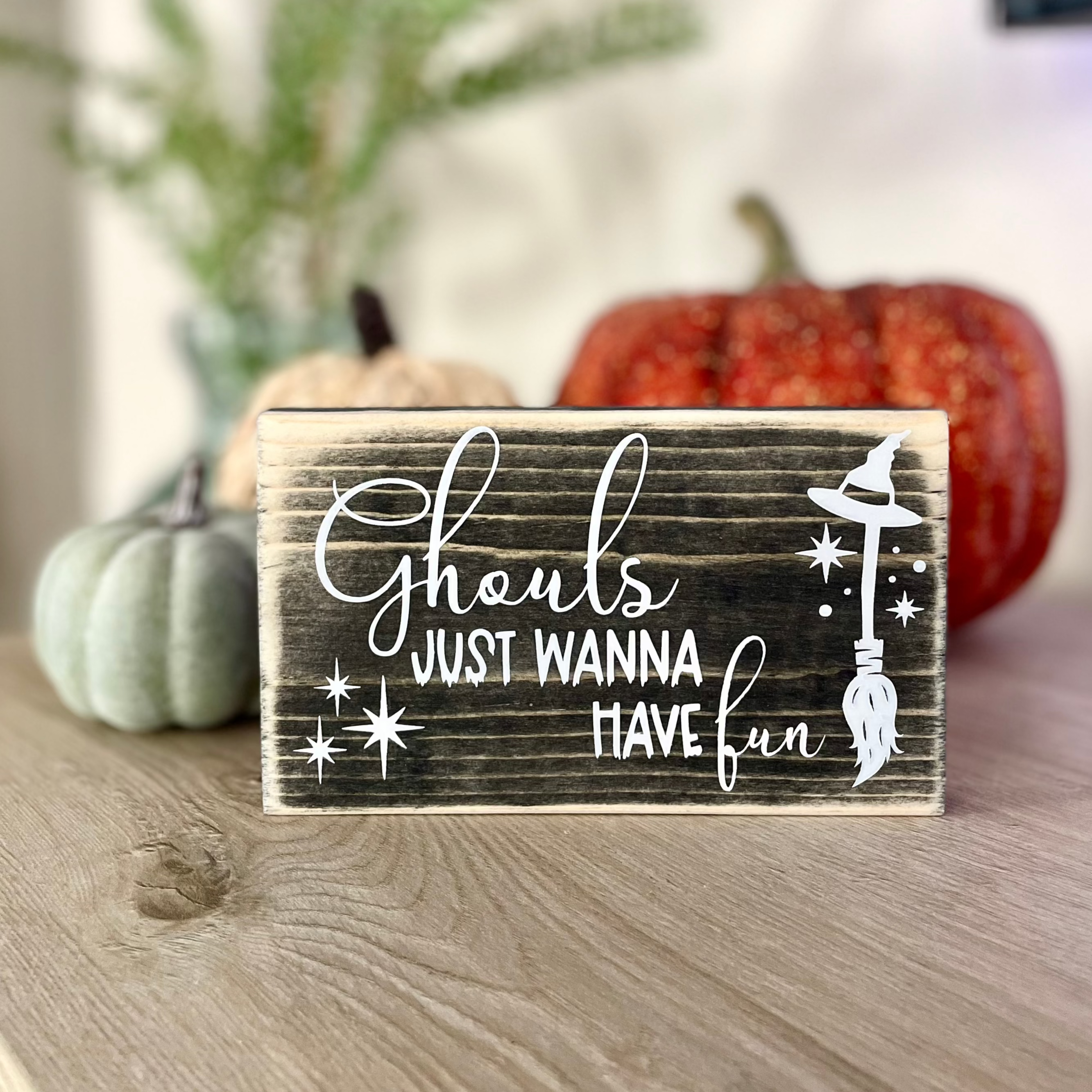 A small, black, wood sign sits on a table with decorative pumpkins.