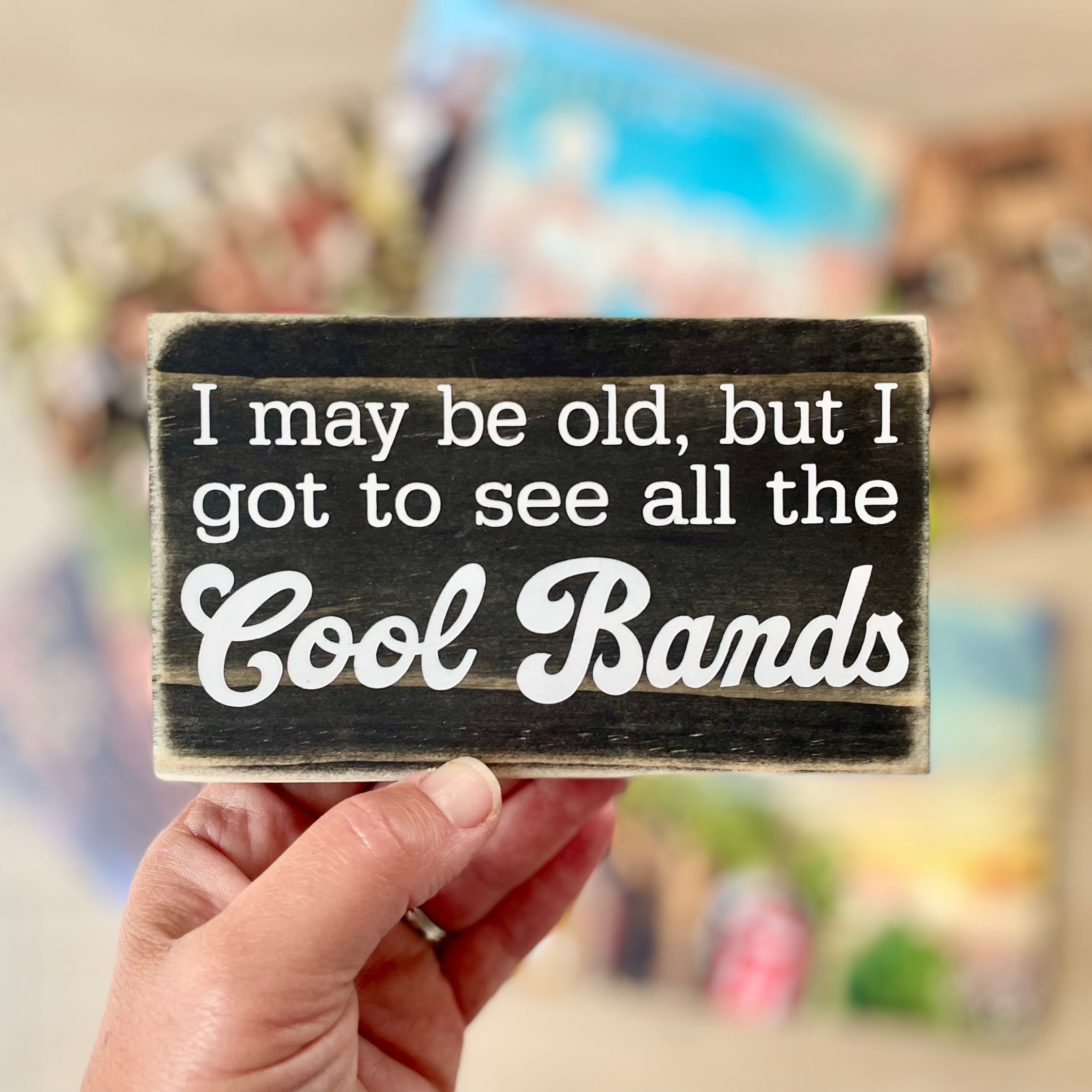 A small, black, wood sign is being held; vinyl records are blurred in the background.