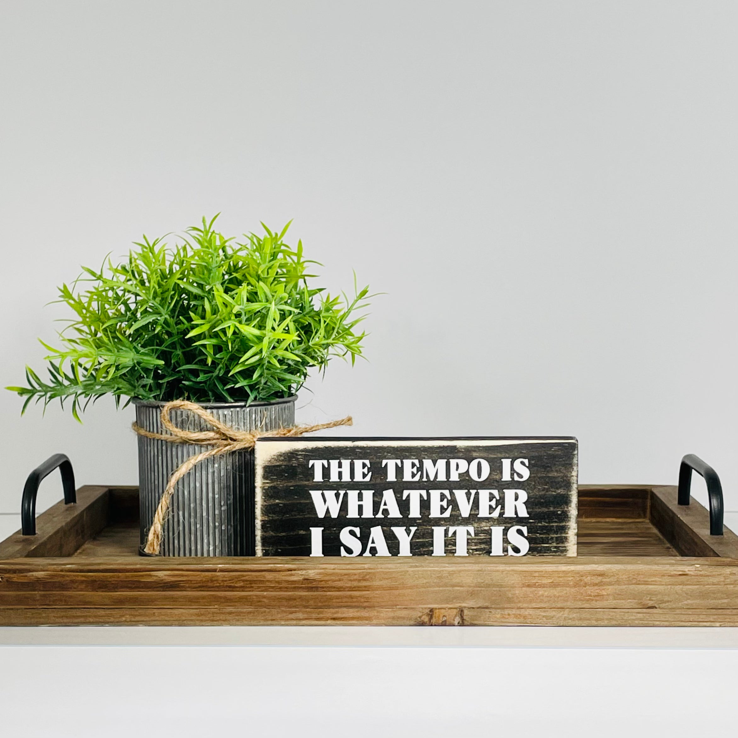 A small, black, wood sign sits on a wood tray with a small plant next to it