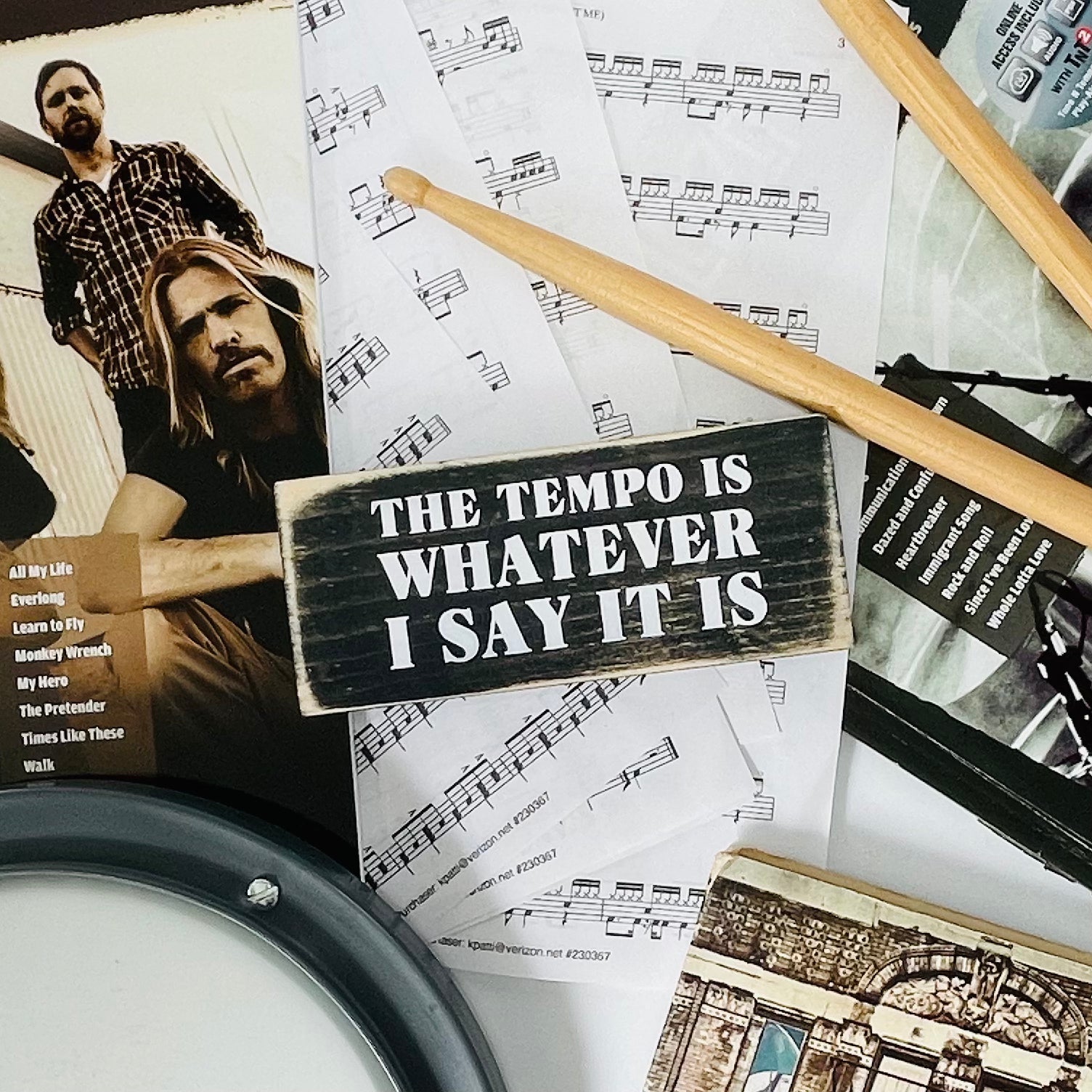 A small, black, wood sign sits on sheet music with drumsticks next to it.
