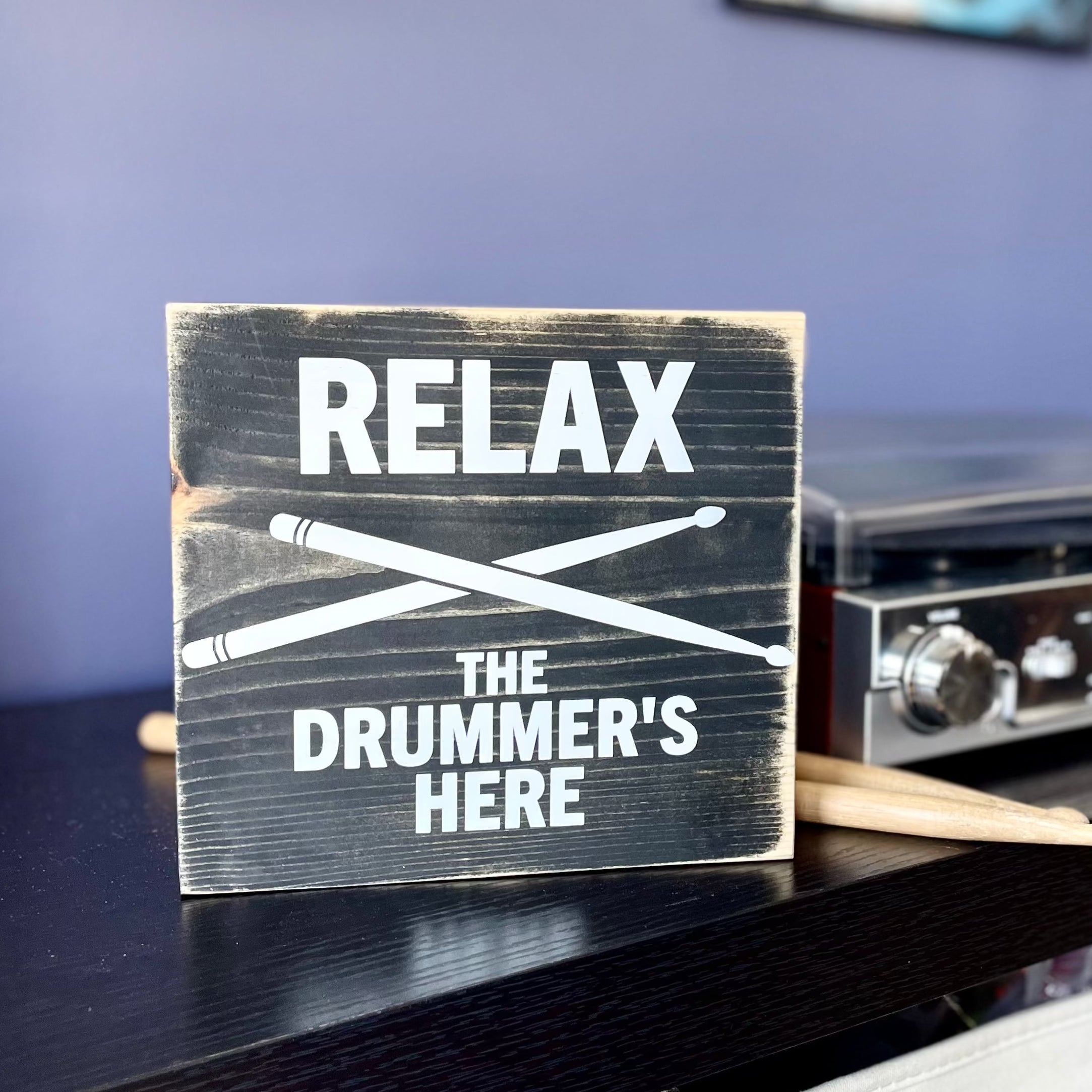 A black, wood sign with white lettering is sitting on a black shelf next to a record player and a set of wood drumsticks. The sign reads: "Relax the Drummer's Here." A white pair of drumsticks is painted in an x under the word relax.