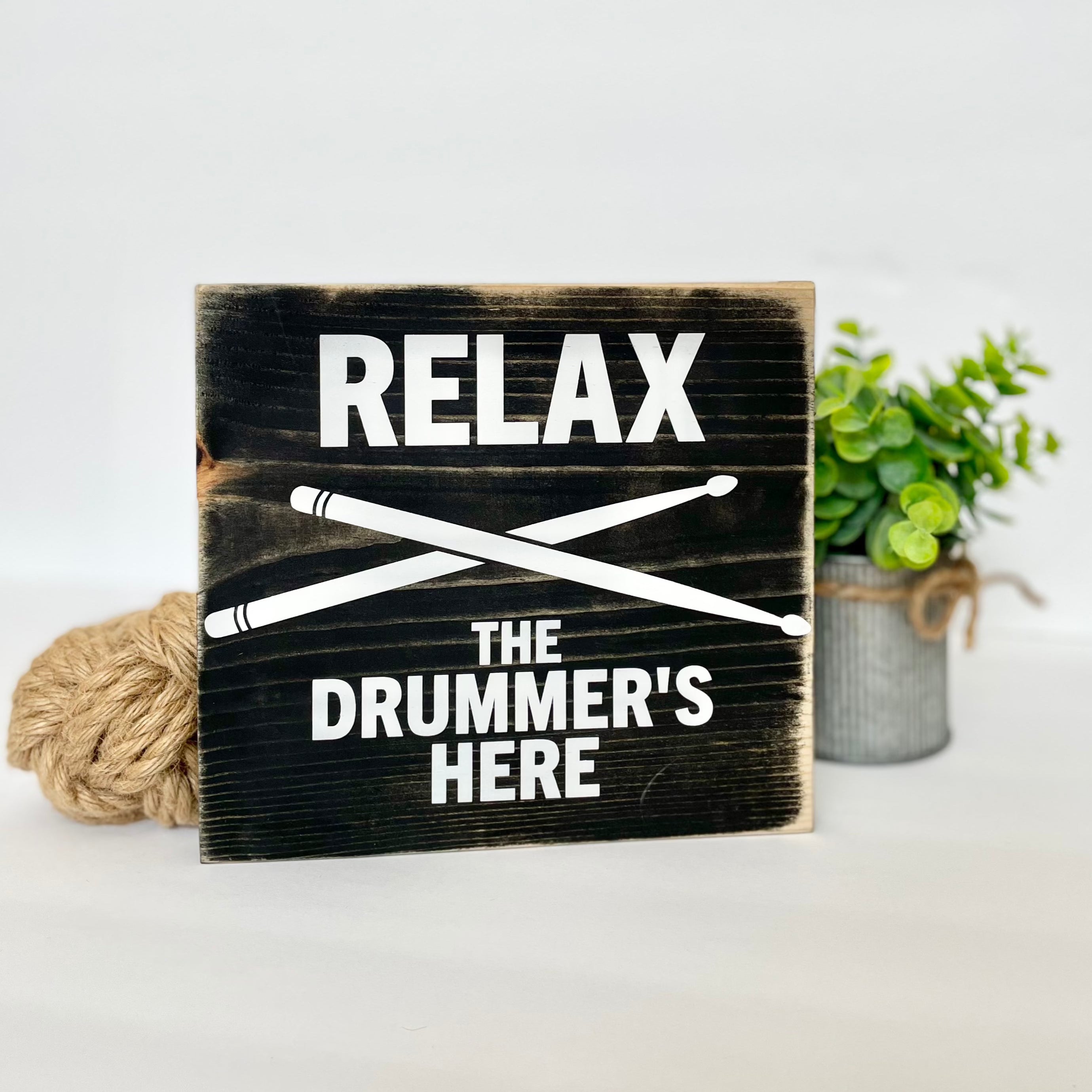 A black, wood sign with white lettering sits on a white shelf next to a small plant. The sign reads: "Relax the Drummer's Here." A white pair of drumsticks is painted in an x under the word relax.