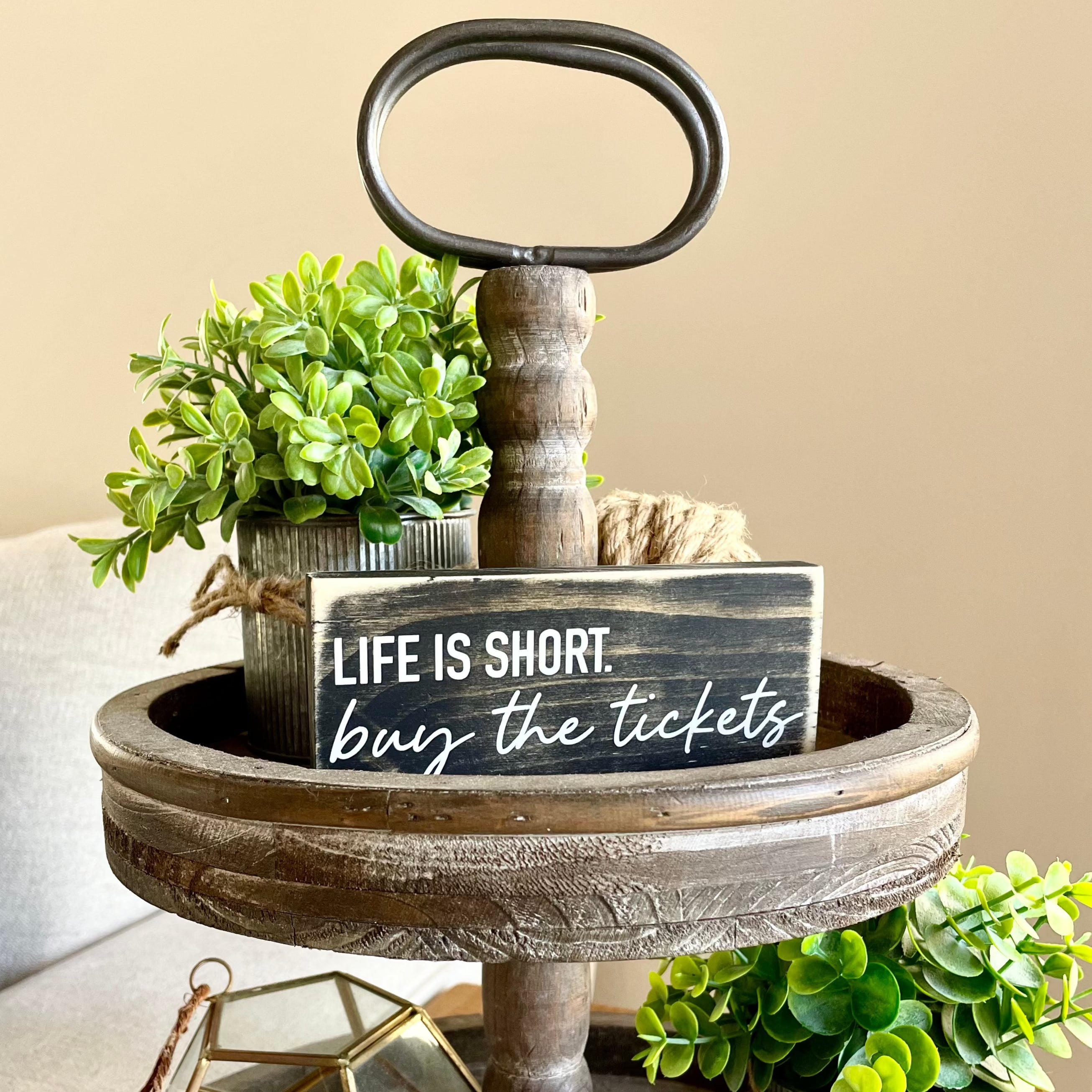 a black sign sits on a tiered tray with a small plant, it says Life is Short. Buy the tickets.