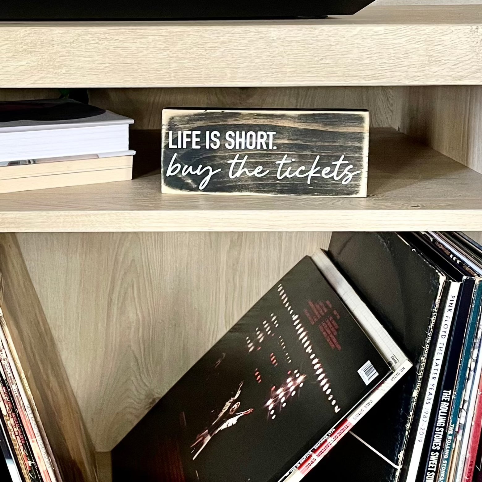 a black sign sits on a shelf with records and books, it says Life is Short. Buy the tickets.