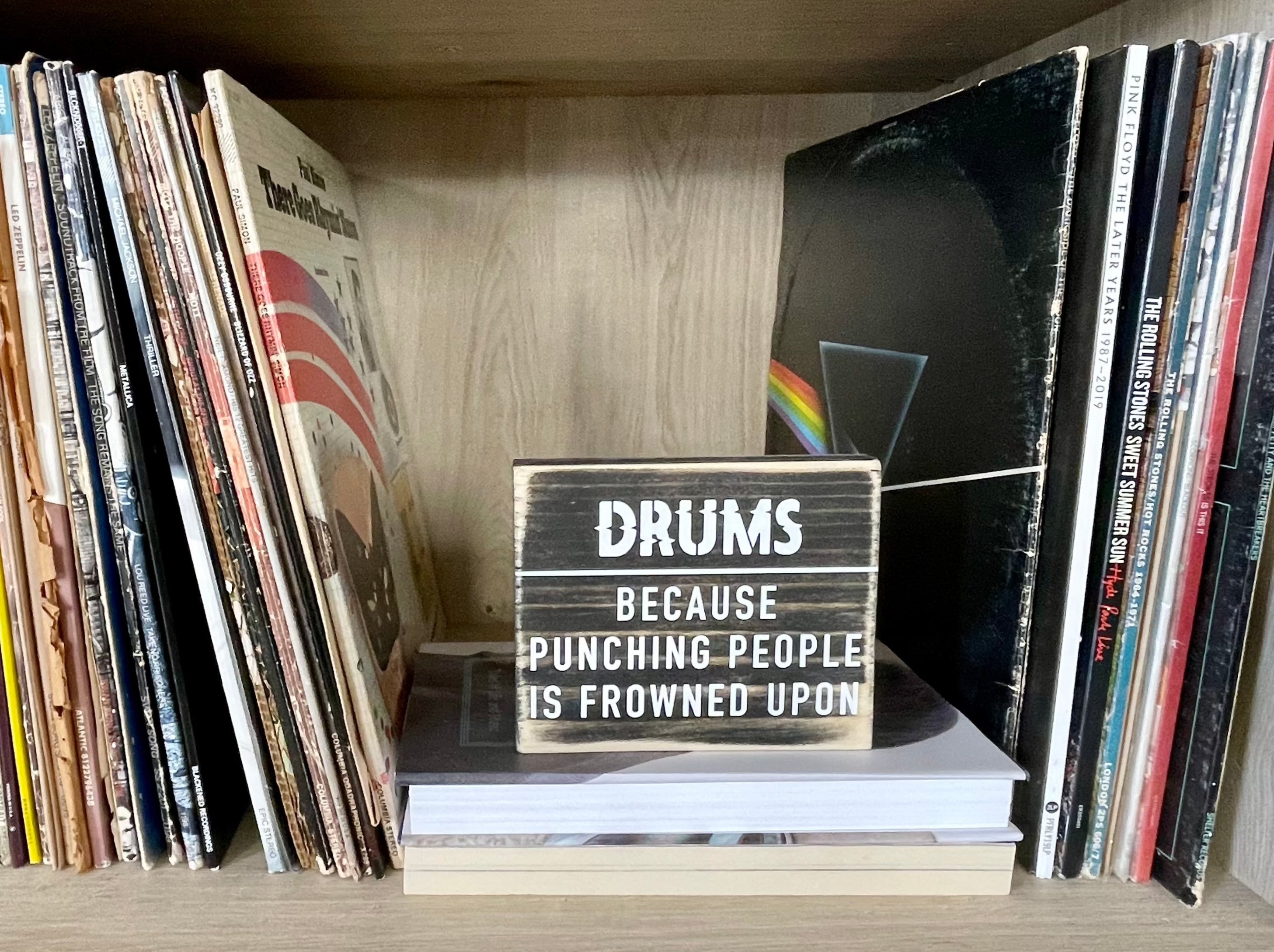 A small, black, wood sign sits on a bookshelf between stacks of vinyl records. In white it reads, "Drums. Because punching people is frowned upon."