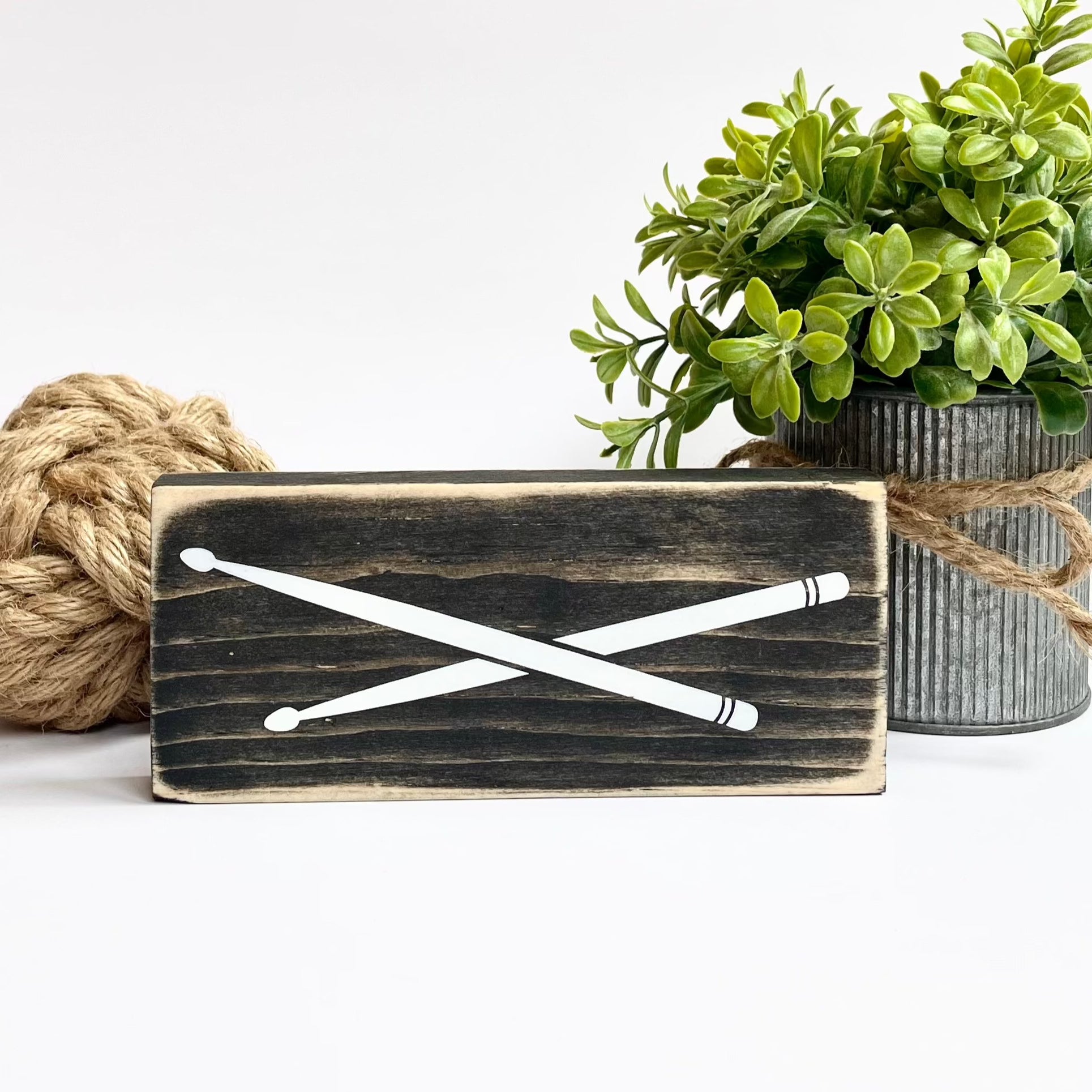 A small, black, wood sign with drumsticks painted on it, sits horizontally on a white surface.