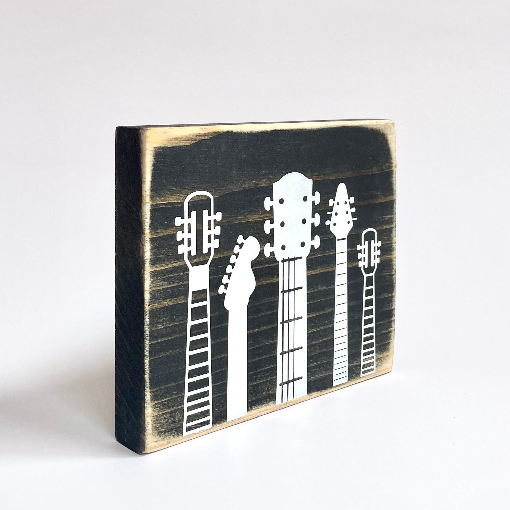 A small, rustic, black, wood sign with white guitar heads on it, sits on a white shelf.