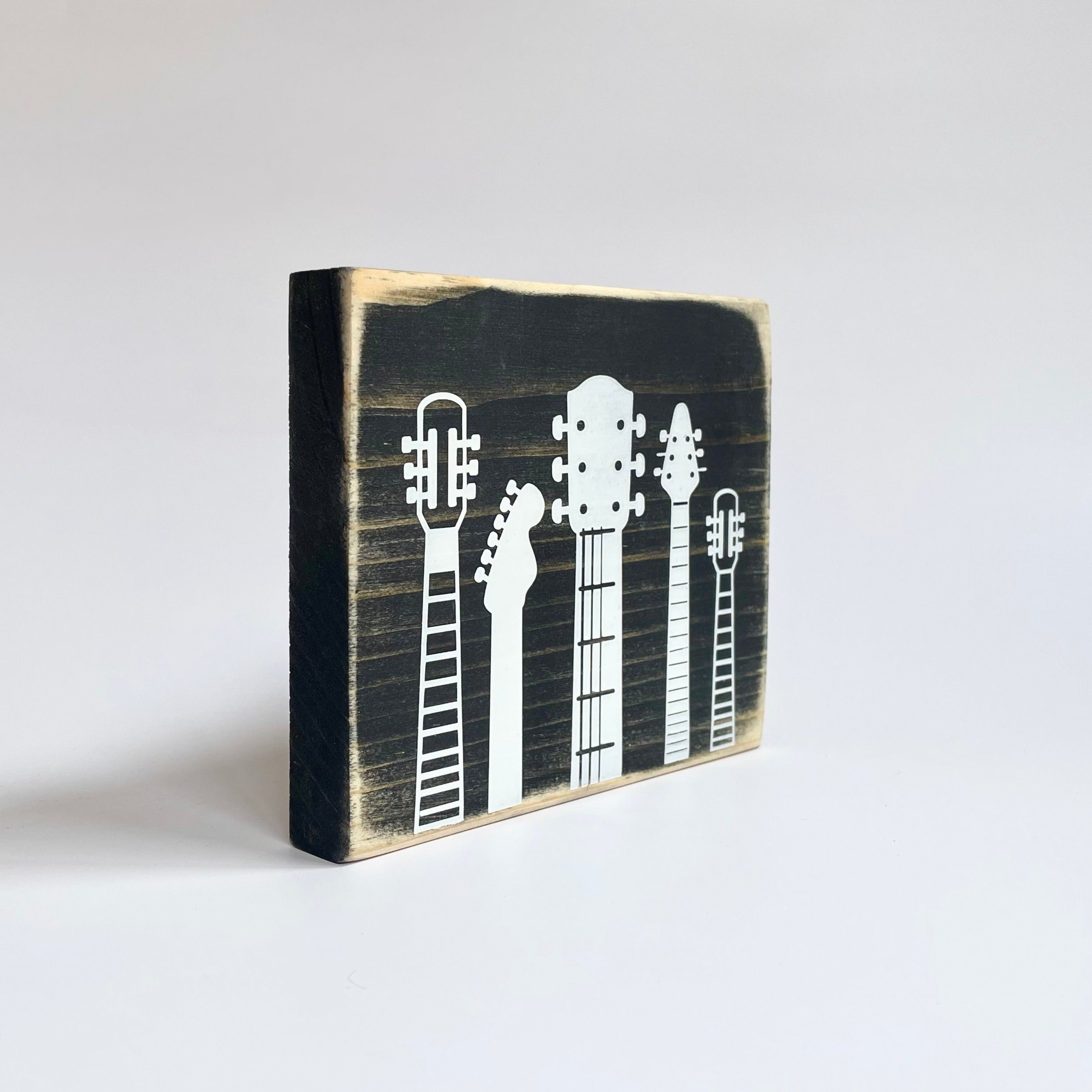 A small, rustic, black, wood sign with white guitar heads on it, sits on a white table..
