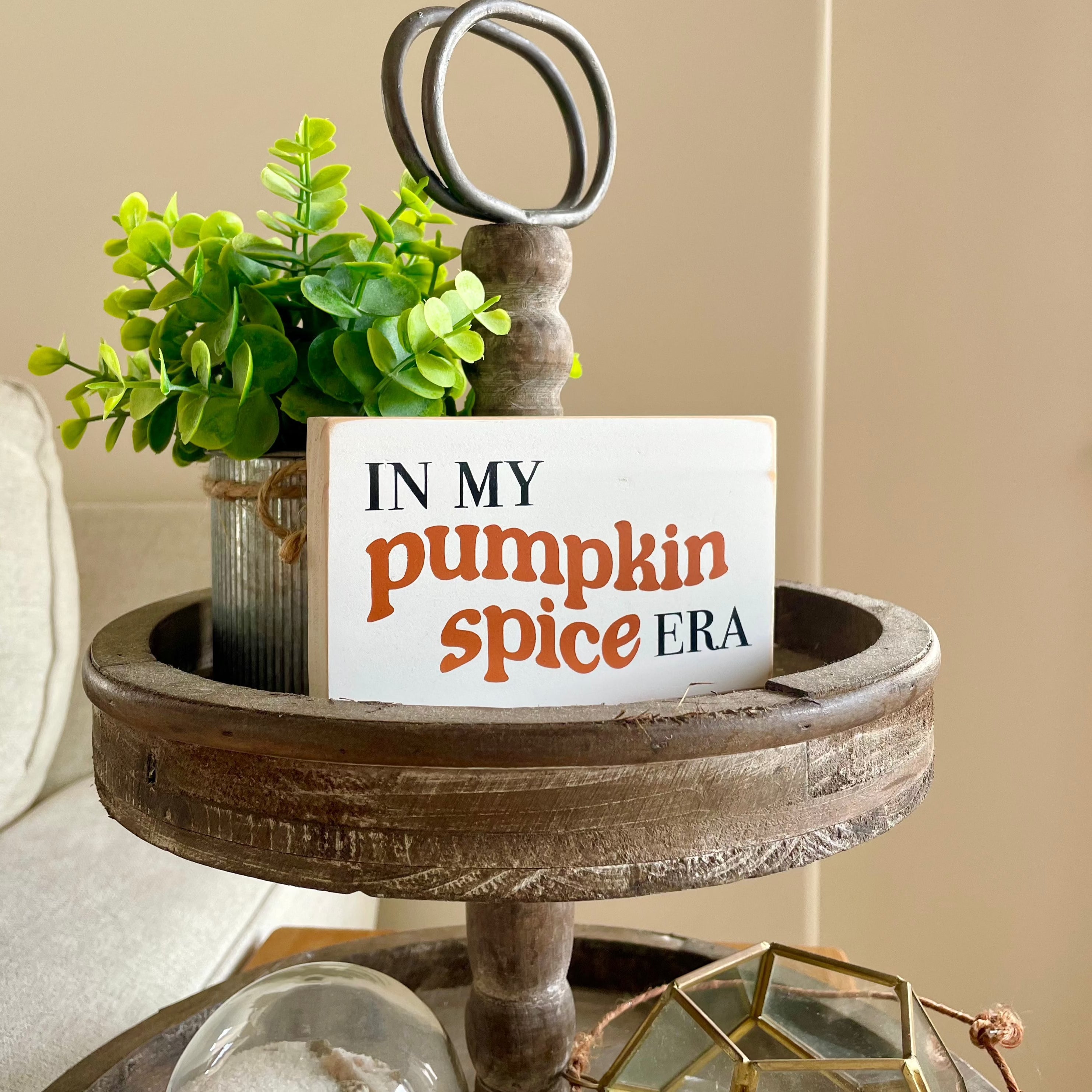 a small, white, wood sign that reads" in my pumpkin spice era" sits on a tiered tray with a plant next to it.