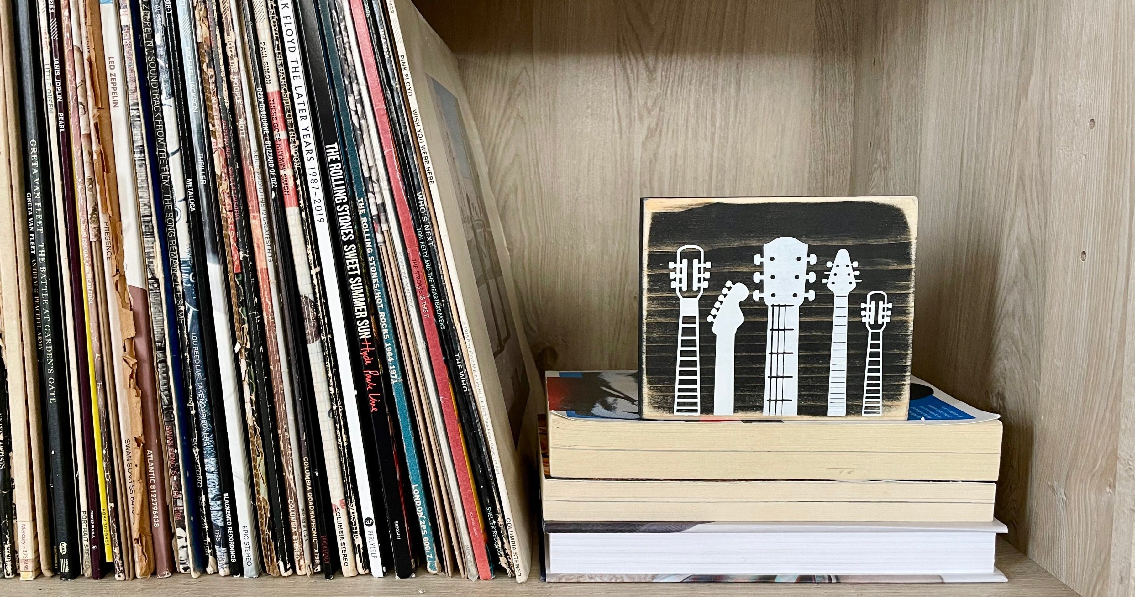 A small, rustic, black, wood sign with white guitar heads on it, sits on a shelf on a stack of books next to vinyl records.