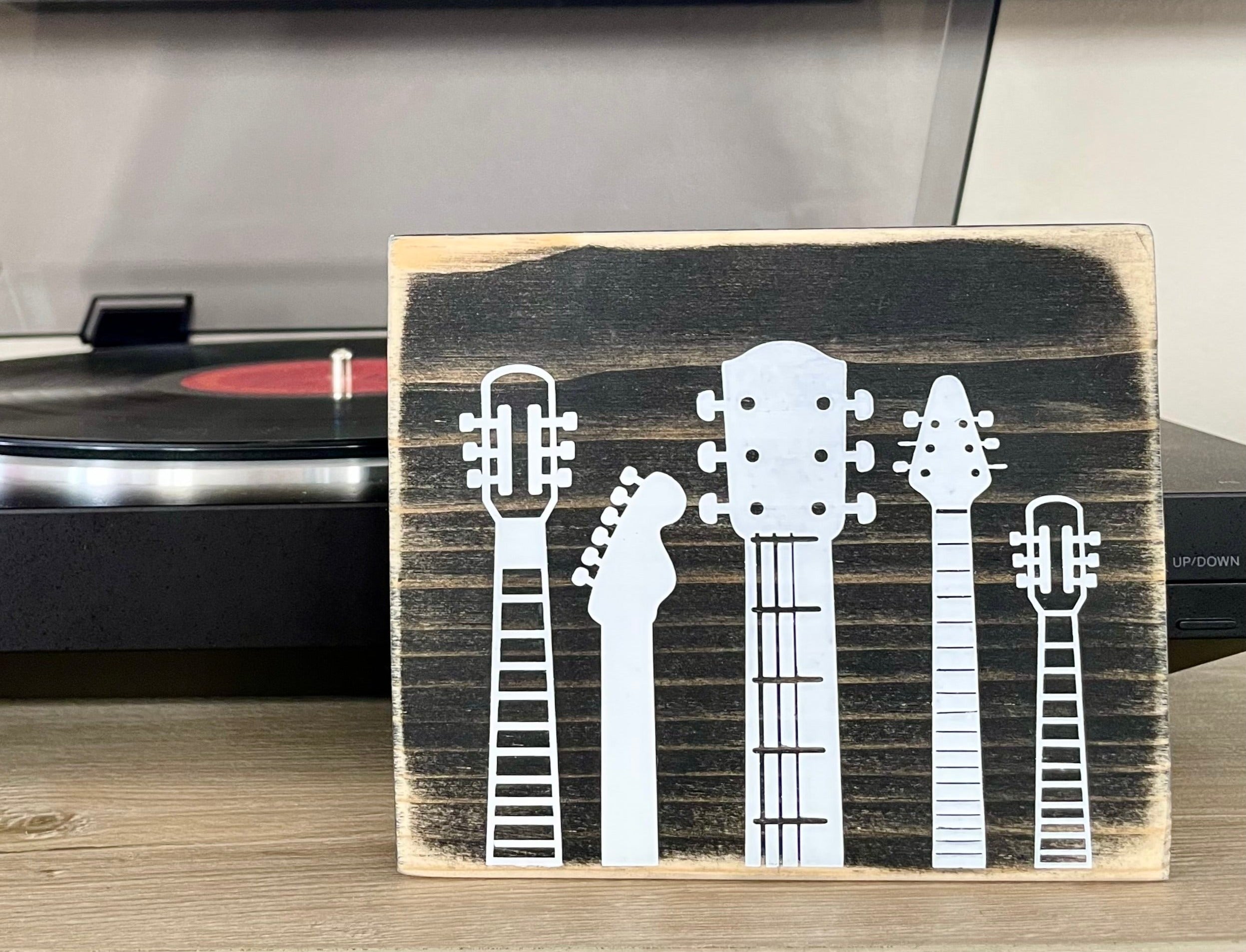 A small, rustic, black, wood sign with white guitar heads on it, sits on a shelf in front of a record player.