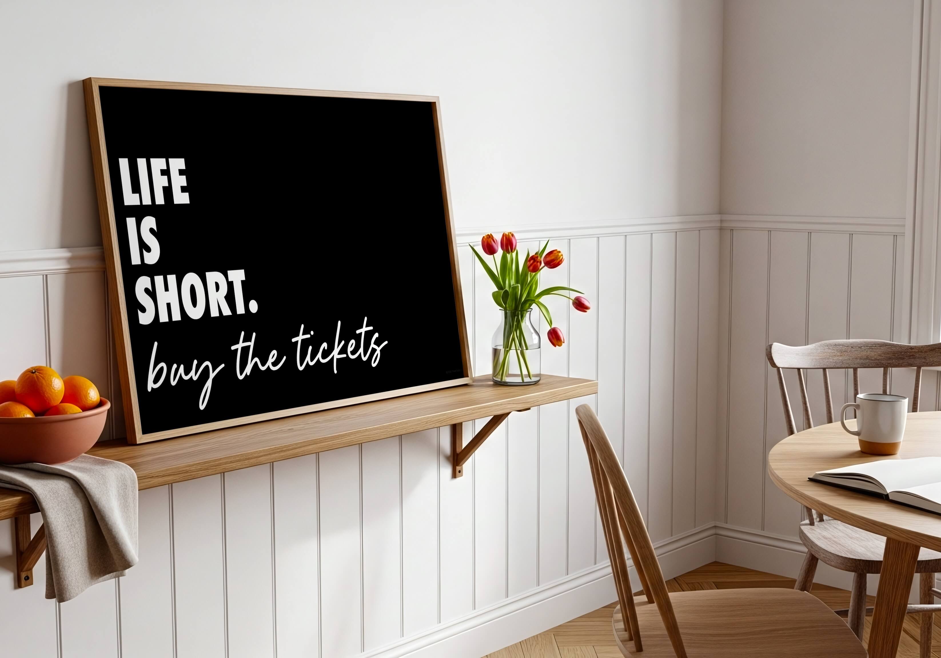 A black framed print with the phrase “LIFE IS SHORT. buy the tickets” in white lettering, leaning on a wooden shelf in a bright dining room with white wainscoting, chairs, a table, and a vase of red tulips.