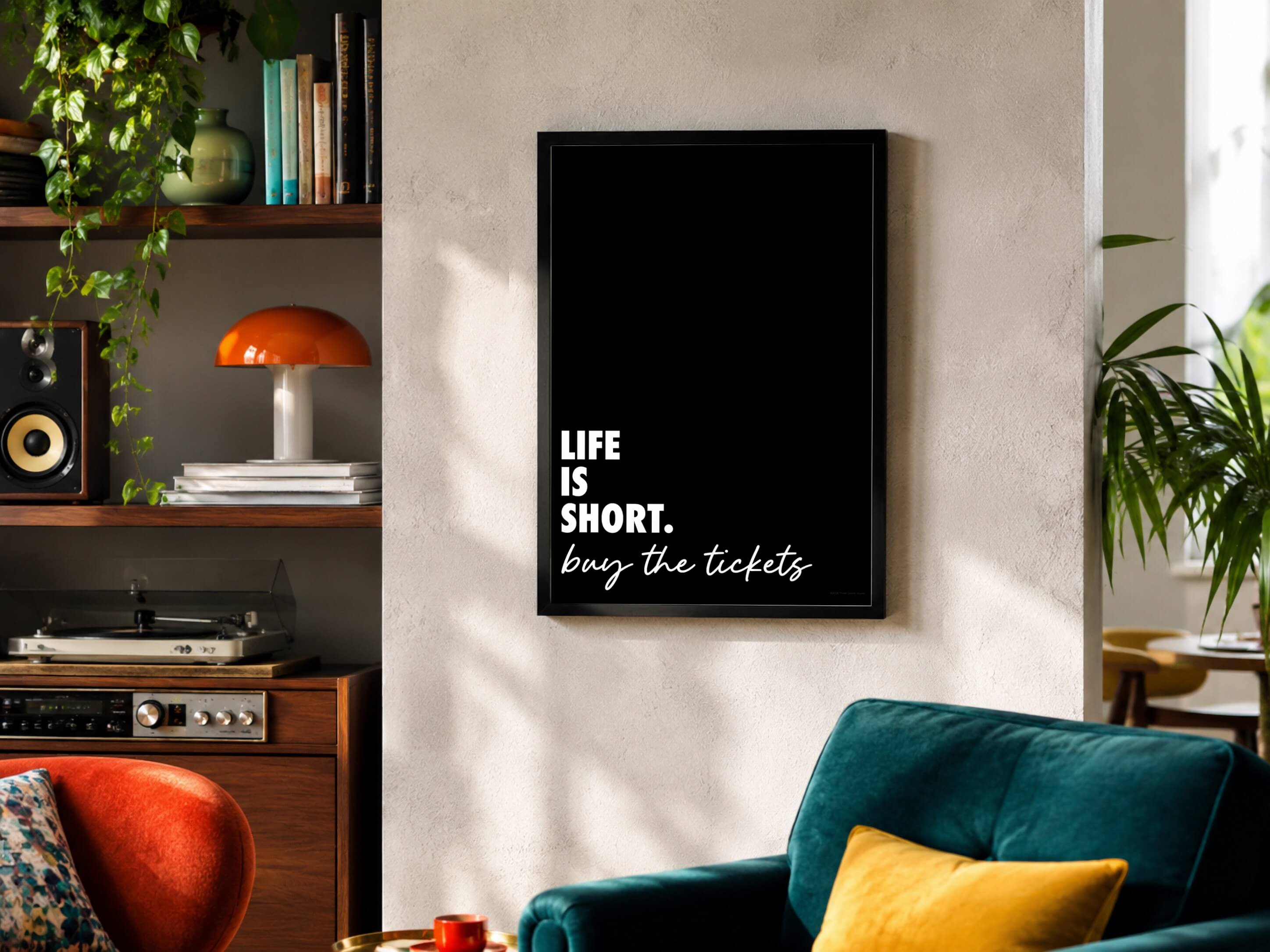 A black framed wall art print with white text reading 'LIFE IS SHORT. buy the tickets' hanging on a textured beige wall in a modern living room with record player on a nearby shelf.