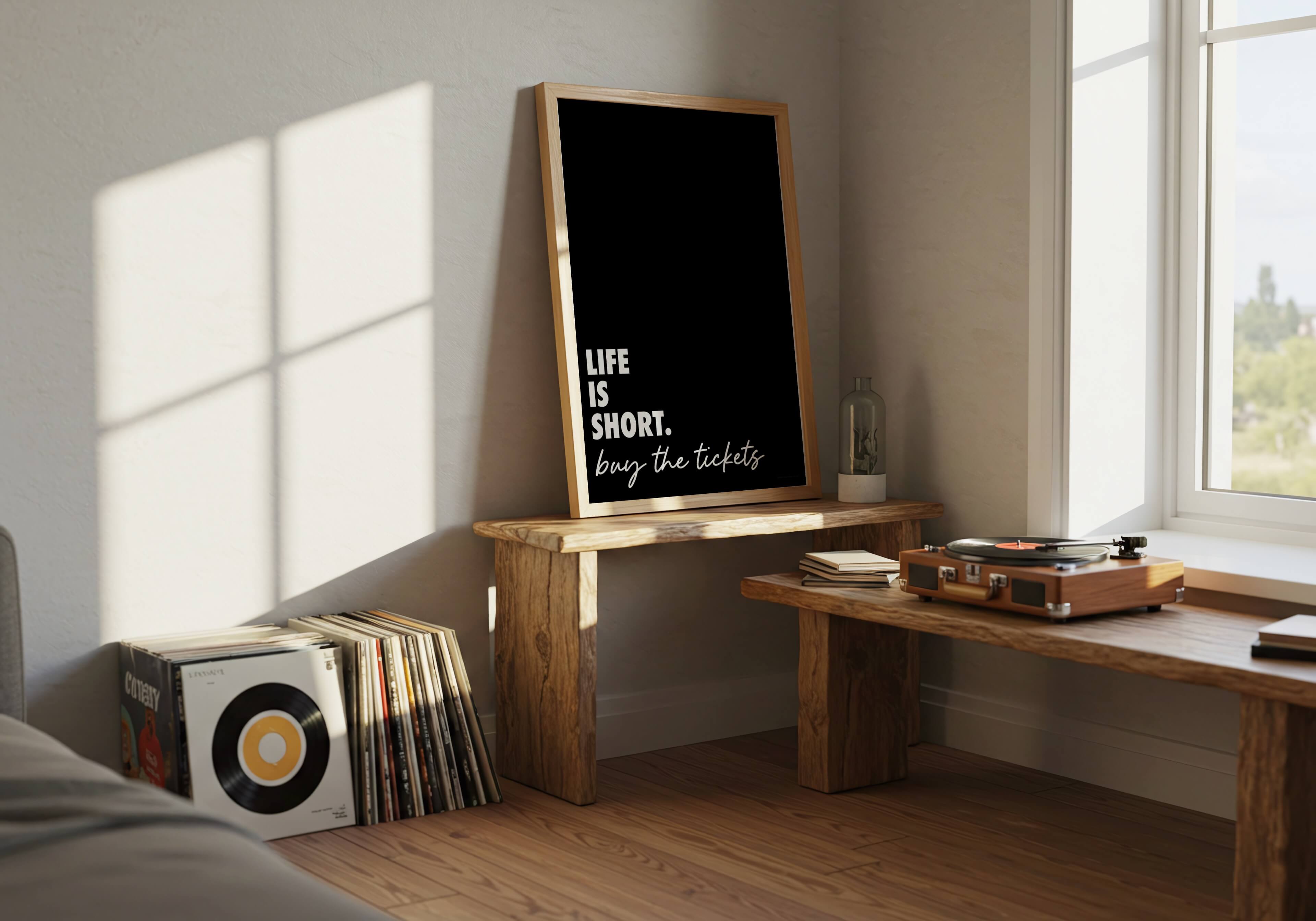 A framed black wall art print with white text reading 'LIFE IS SHORT. buy the tickets leans against a wall near records and a record player.