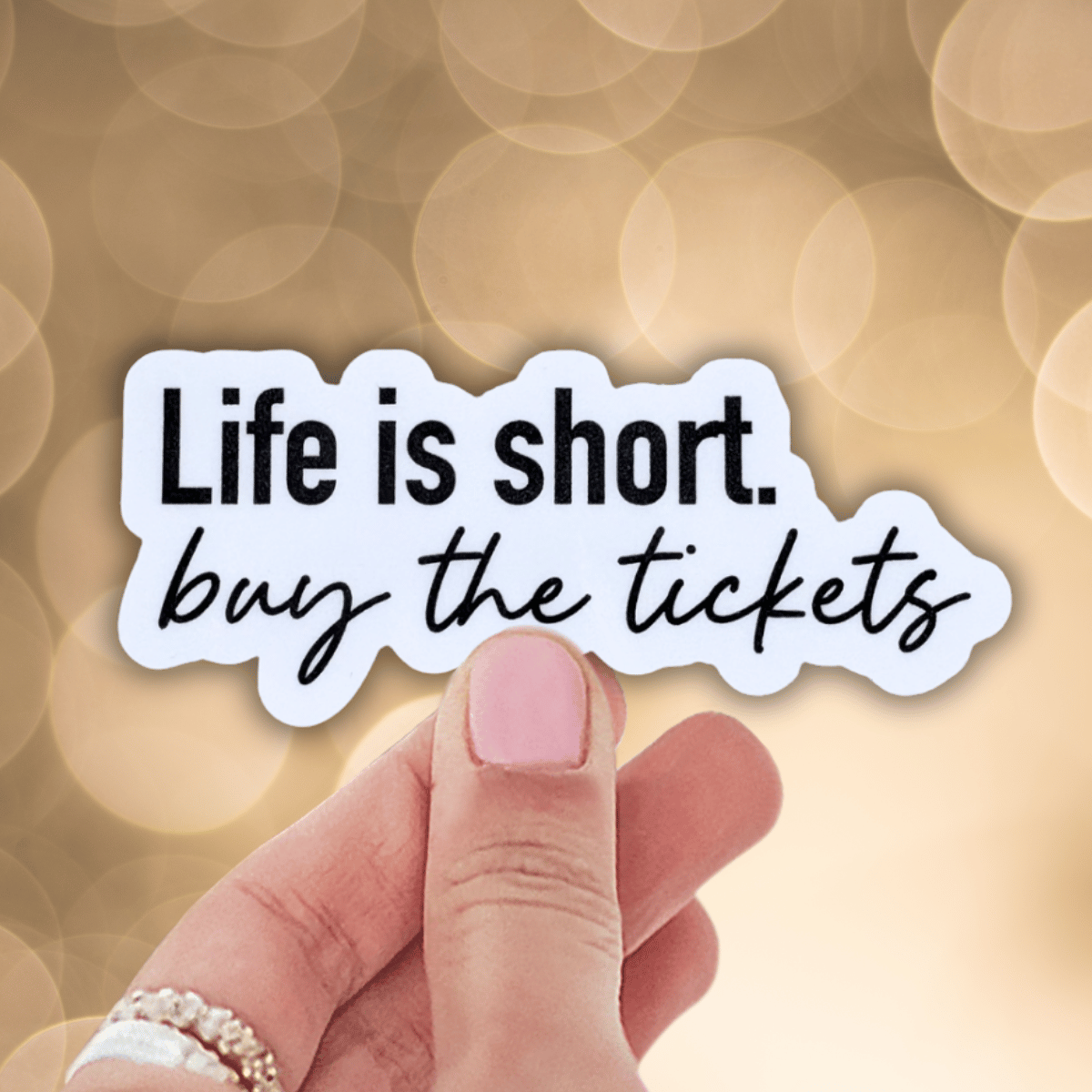 Hand holding a sticker with 'Life is short, buy the tickets' text against a blurred background.