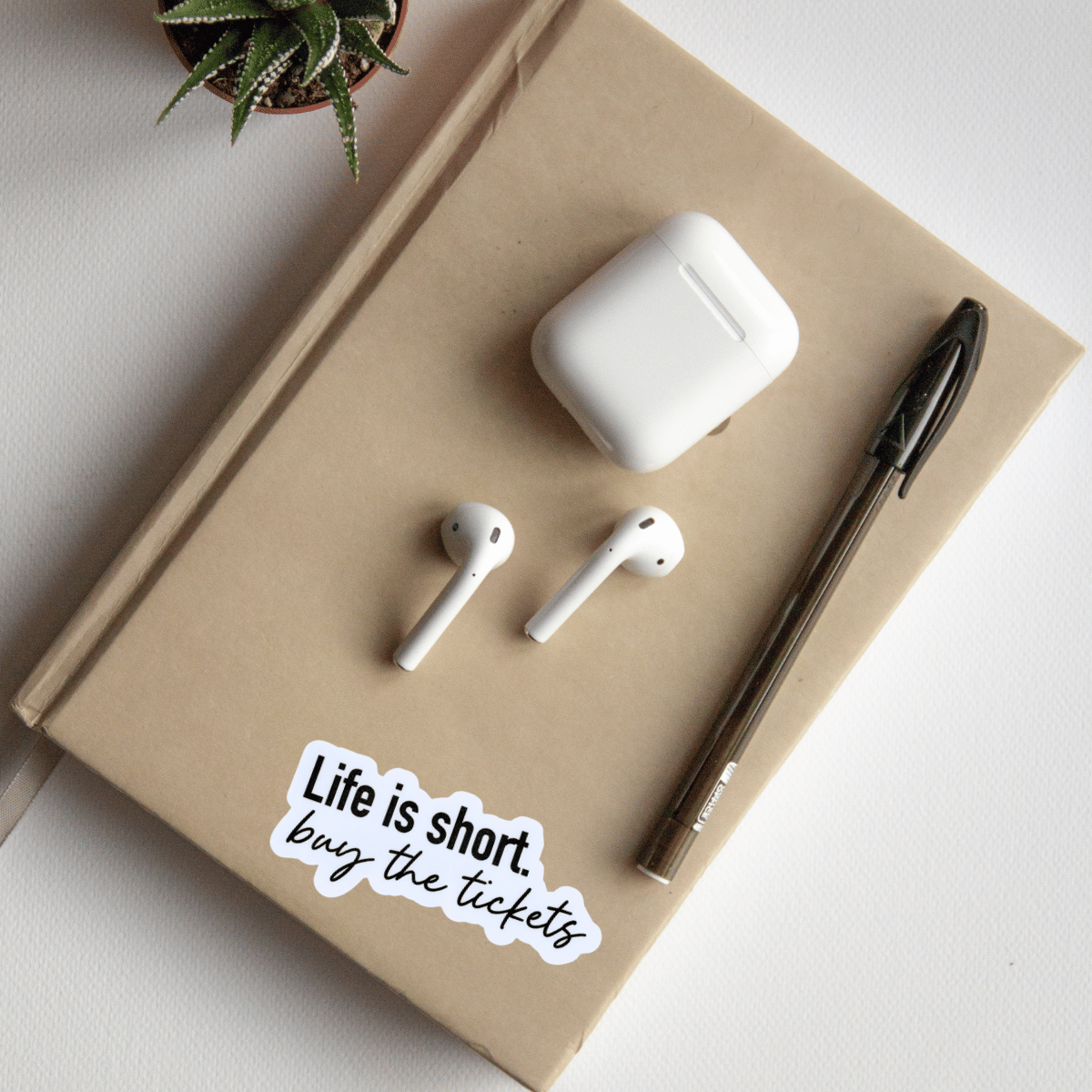 White AirPods, pen, and 'Life is short, buy the tickets' sticker on a beige notebook.