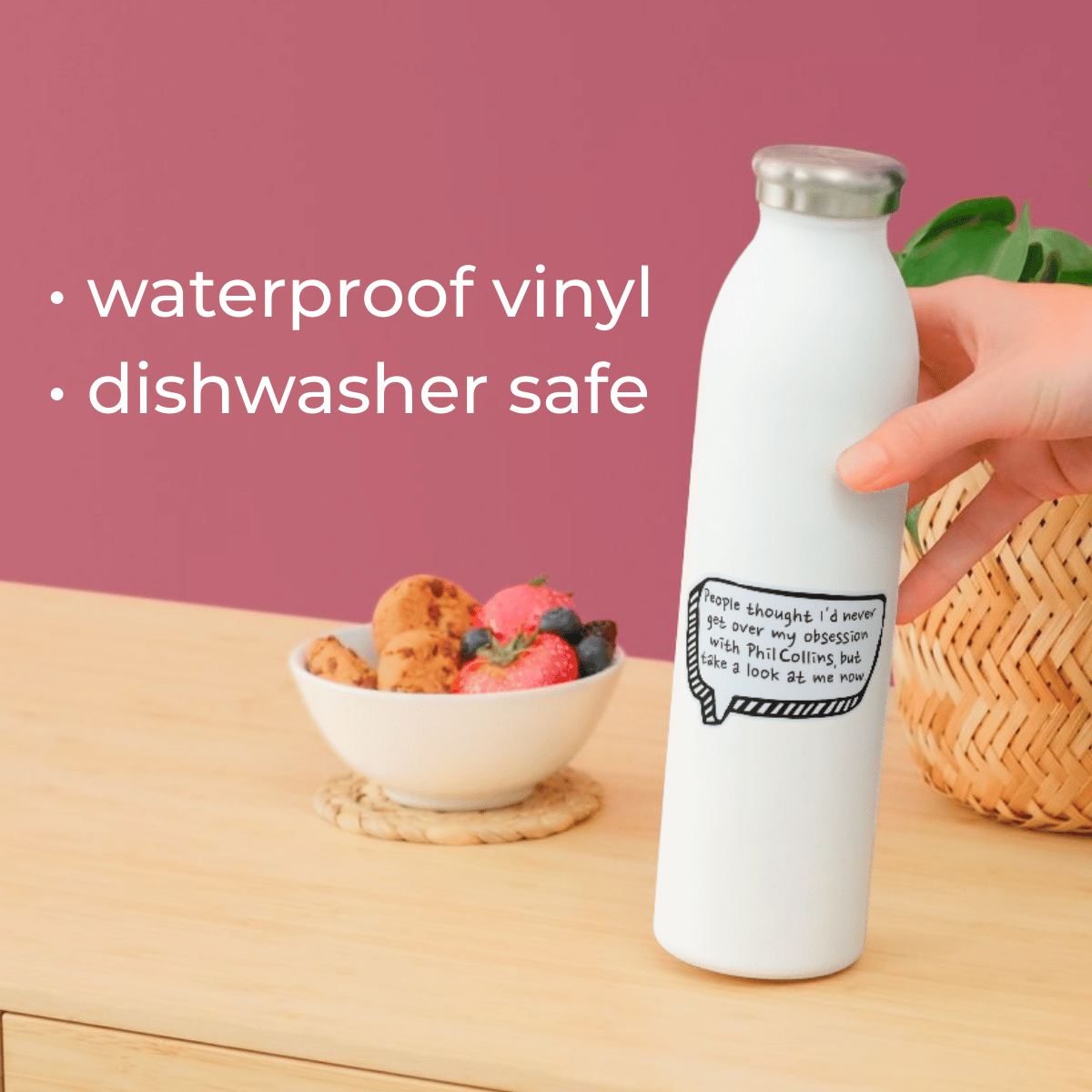 White water bottle with text on a wooden table with a bowl of fruit and a pink background.