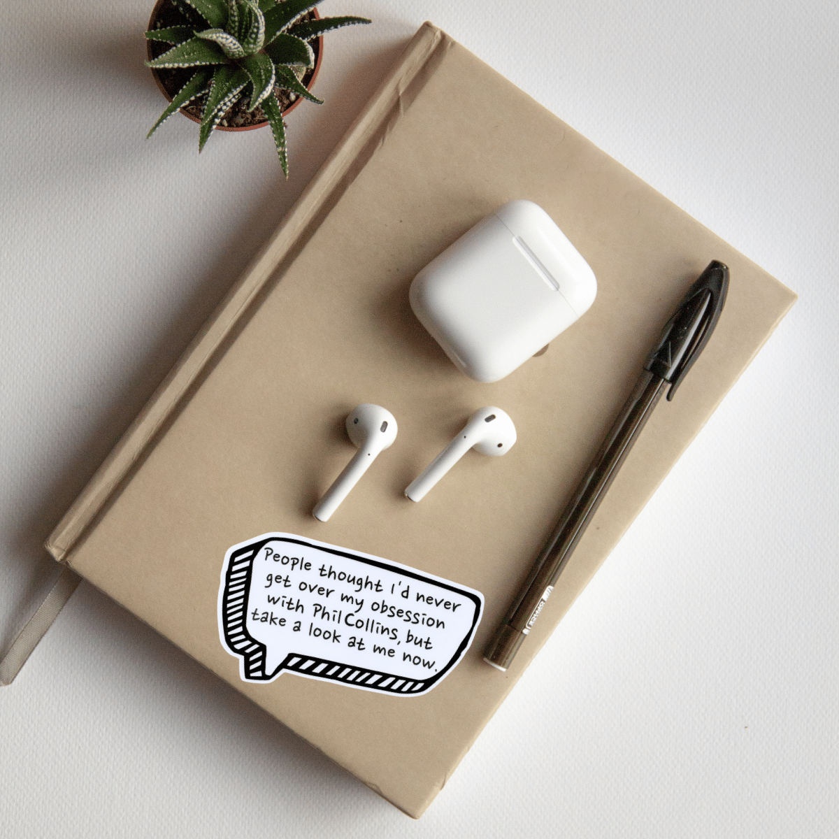 Notebook with AirPods case, AirPods, and a pen on a light surface with a plant in the corner.