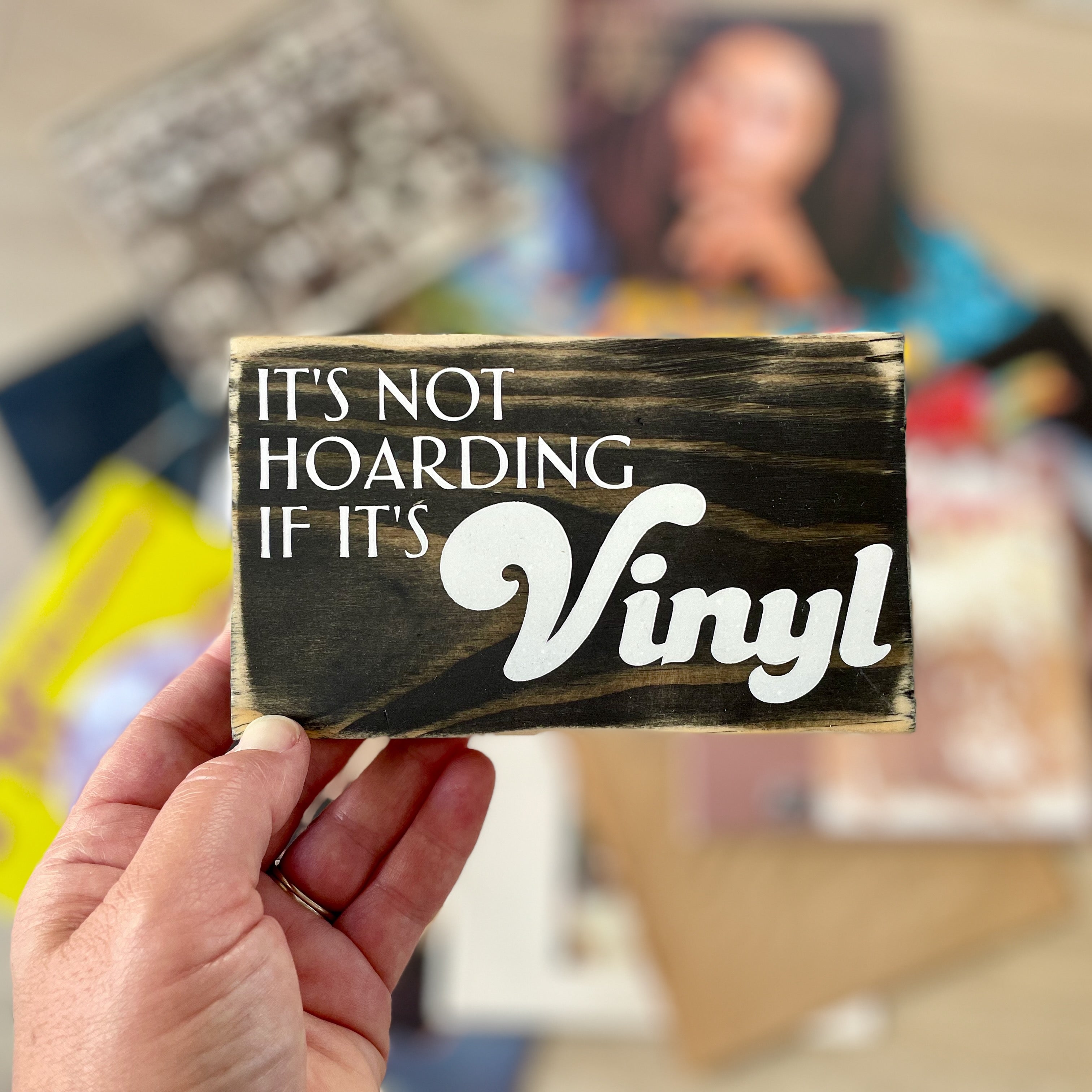 A small, black, wood sign is being held in someone's hand, vinyl record jackets are blurry in the background. In white, the sign reads, "It's not hoarding if it's vinyl".