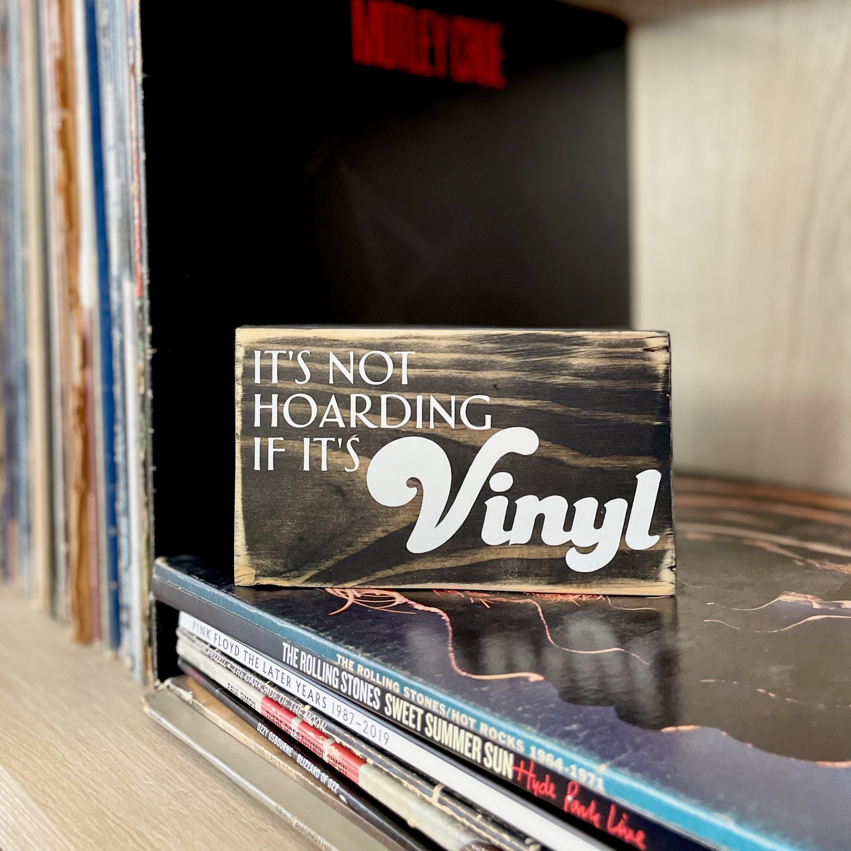 A small, black, wood sign sits on a stack of records on a shelf. In white, the sign reads, "It's not hoarding if it's vinyl".
