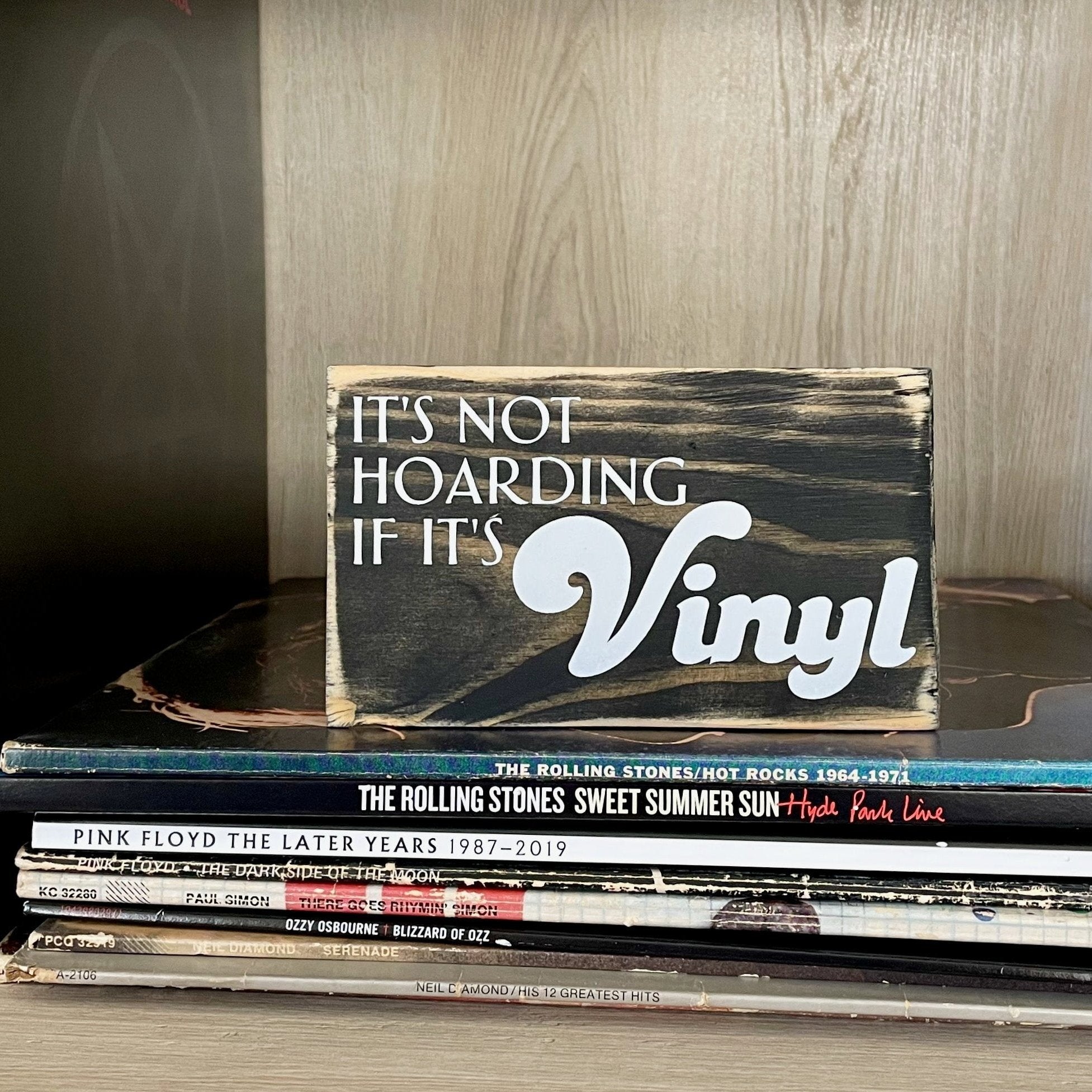 A small, black, wood sign sits on a stack of records on a shelf full of records. In white, the sign reads, "It's not hoarding if it's vinyl".