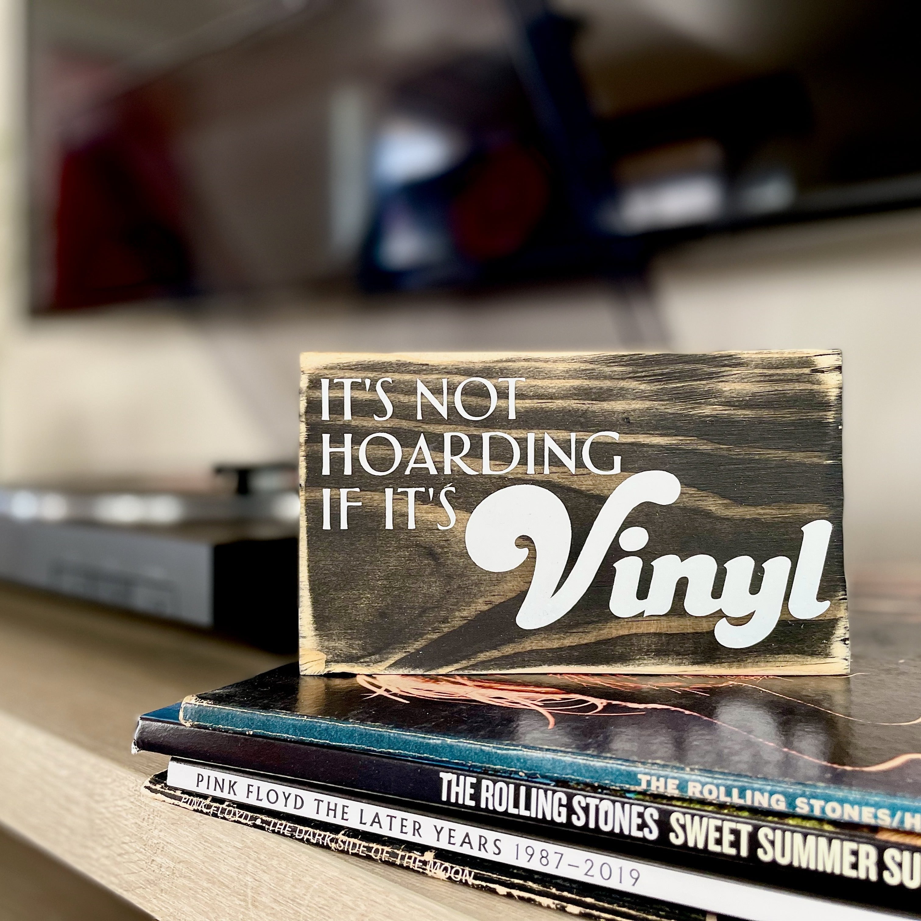 A small, black, wood sign sits on a stack of records next to a record player. In white, the sign reads, "It's not hoarding if it's vinyl".