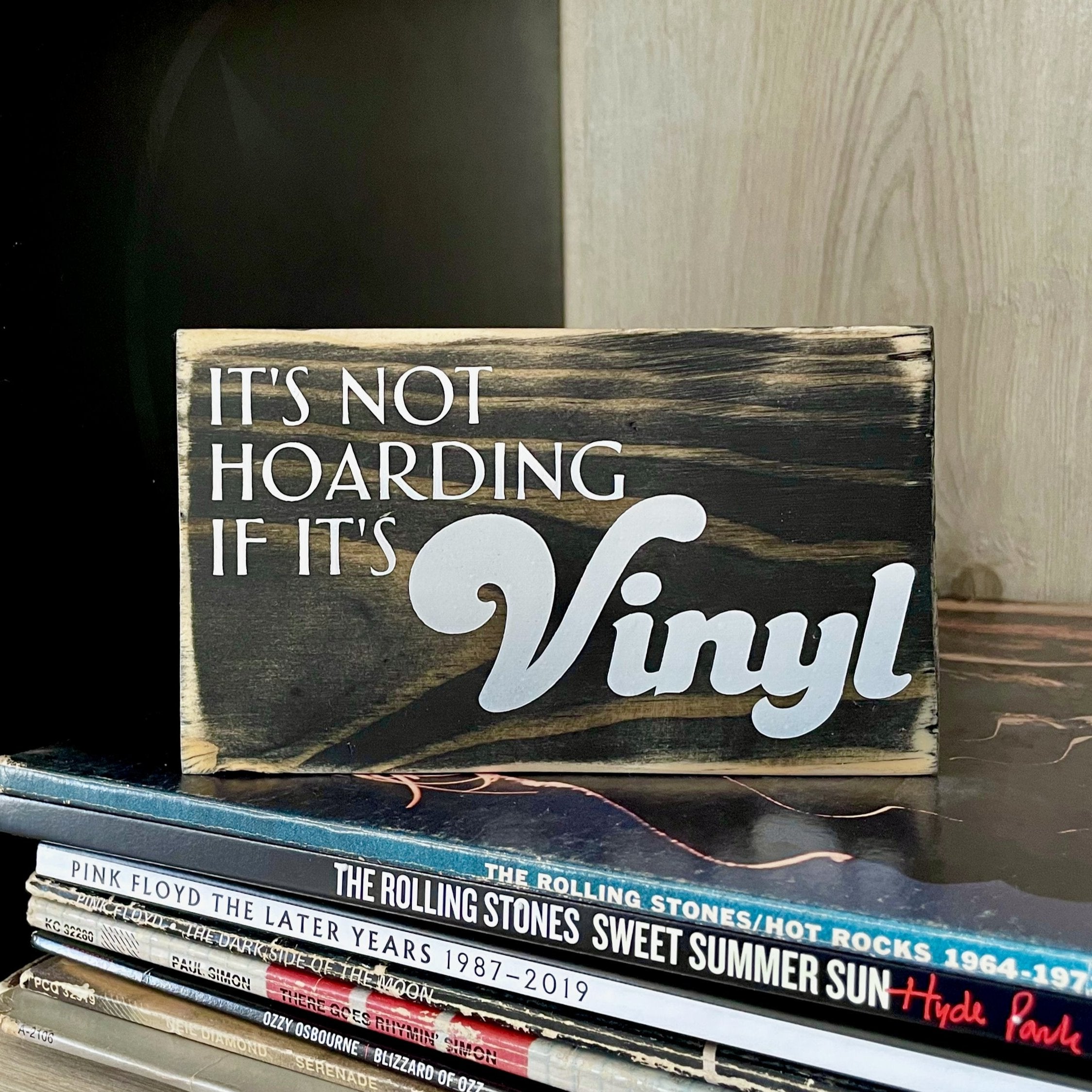 A close up of a small, black, wood sign sitting on a stack of records on a shelf. In white, the sign reads, "It's not hoarding if it's vinyl".