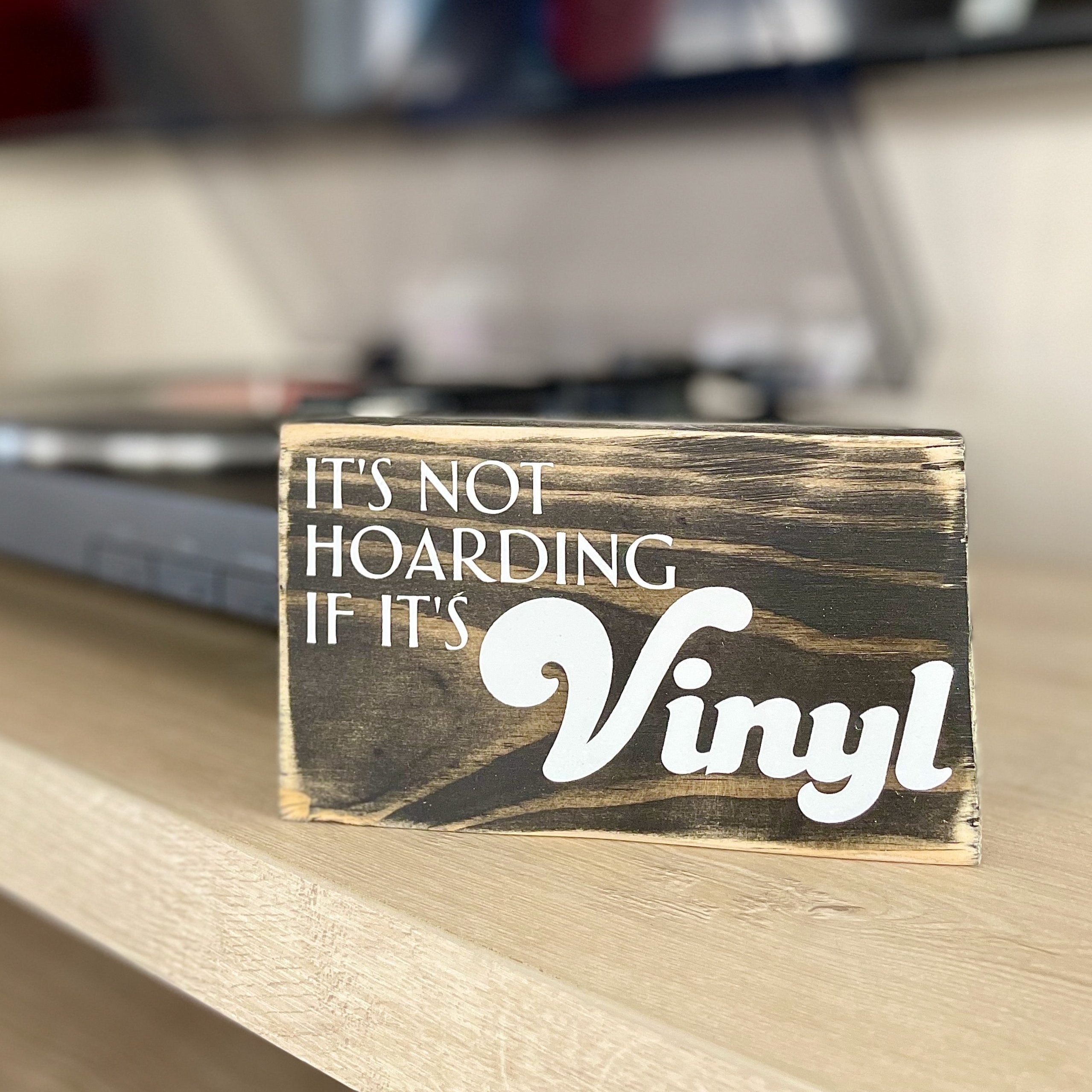 A small, black, wood sign sits on a table next to a record player. In white, the sign reads, "It's not hoarding if it's vinyl".