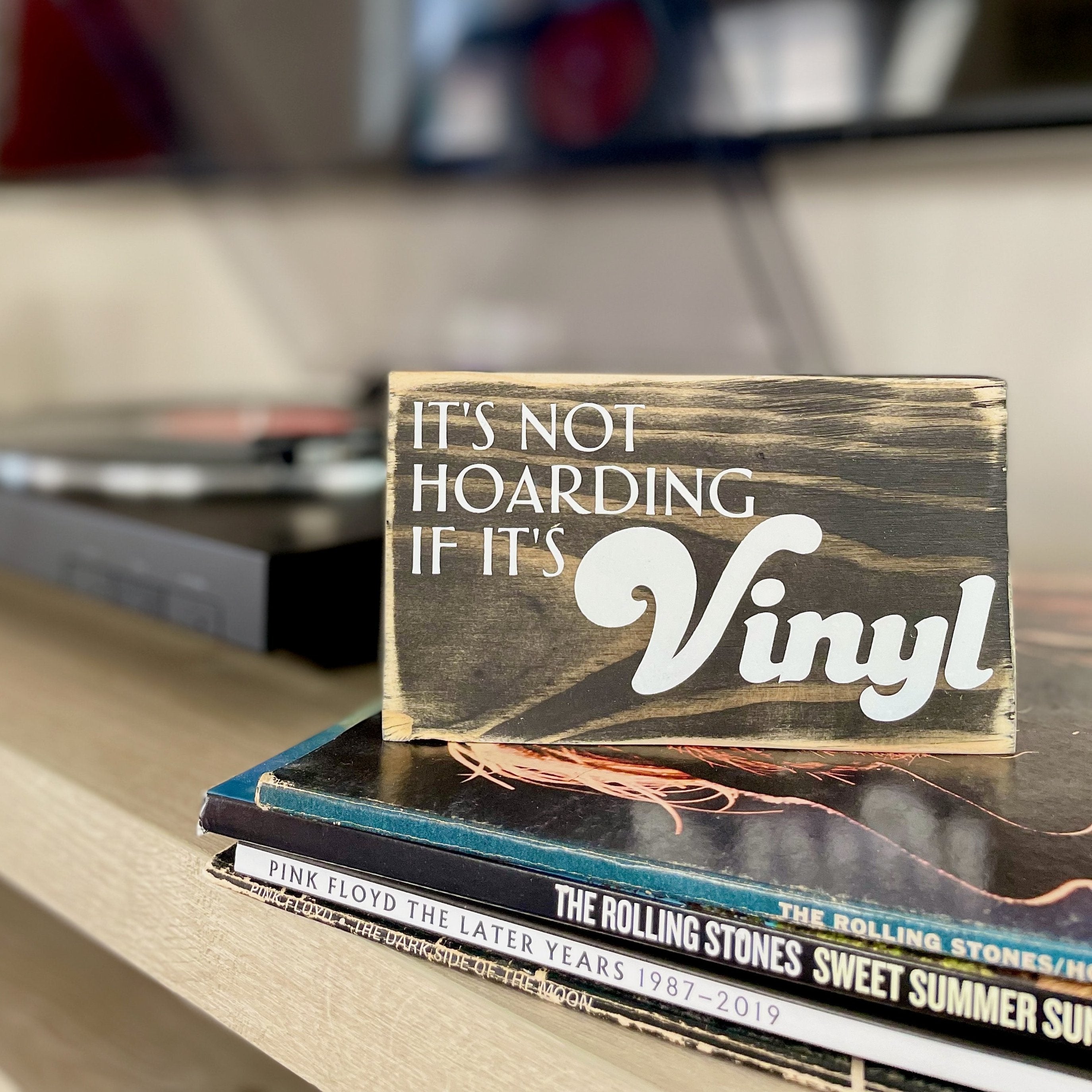 A small, black, wood sign sits on a stack of records on a shelf next to a record player. In white, the sign reads, "It's not hoarding if it's vinyl".