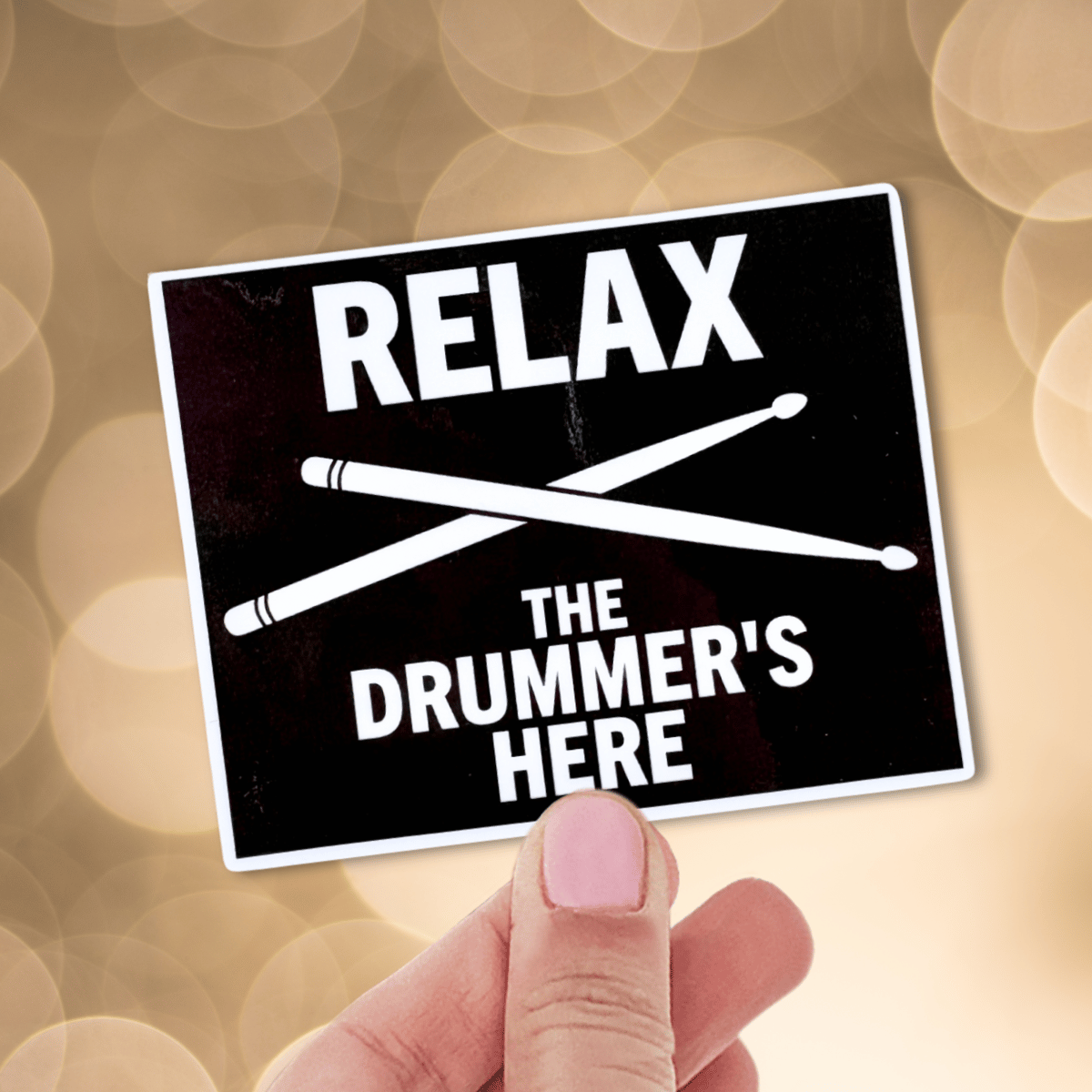 Sticker with 'Relax, the drummer's here' text held by a hand against a blurred background