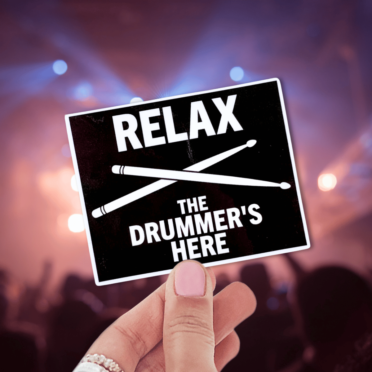 Hand holding a sign with 'Relax, the drummer's here' text against a concert background.