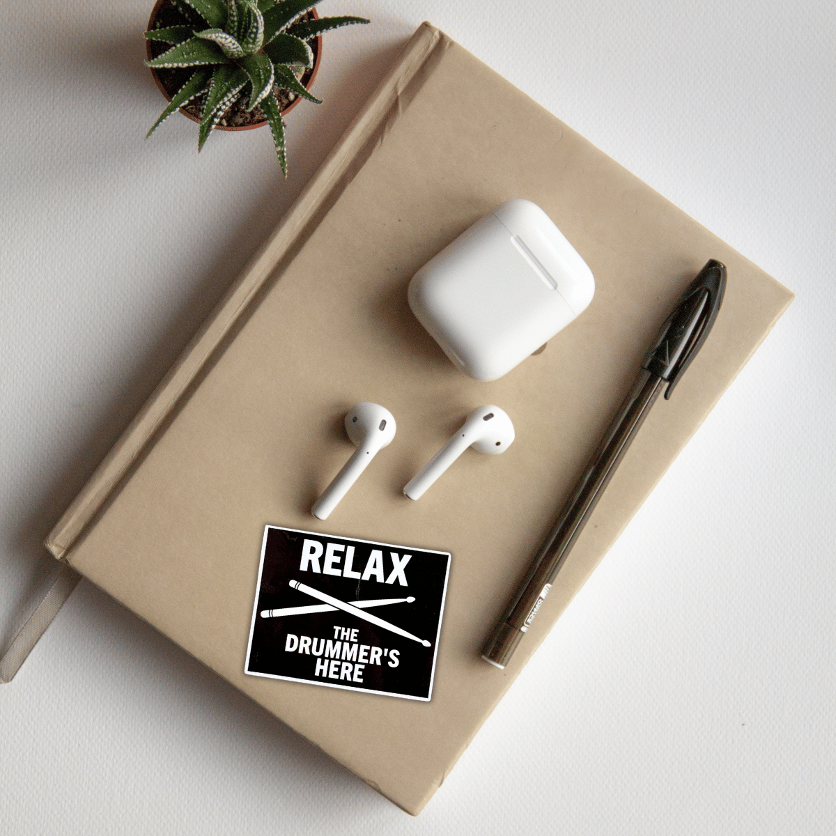Notebook with white earbuds, charging case, pen, and 'Relax the Drummer's Here' sticker on a light background