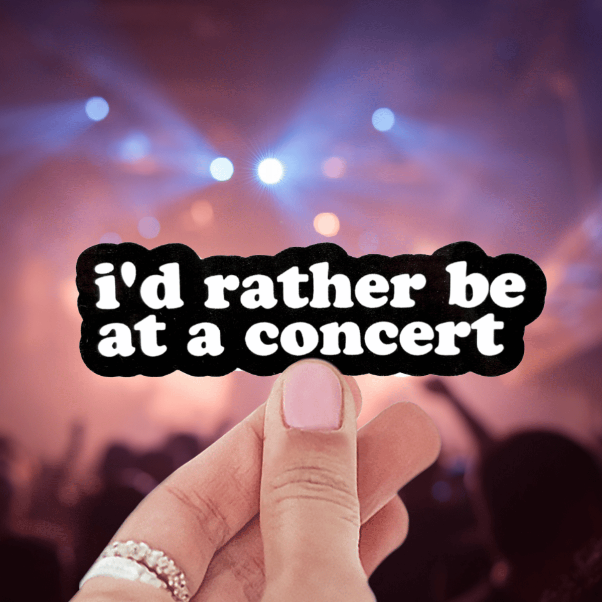 Hand holding a sticker with 'I'd rather be at a concert' text against a concert background.