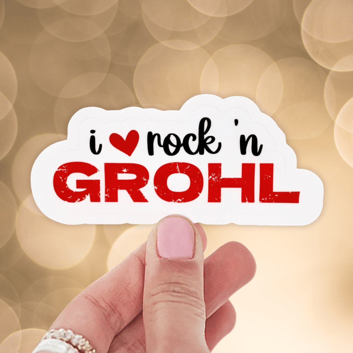 A person is holding a sticker that says "I love Rock n Grohl" on a blurred, gold background. The word Grohl is in a red distressed font and a red heart replaces the word "love"