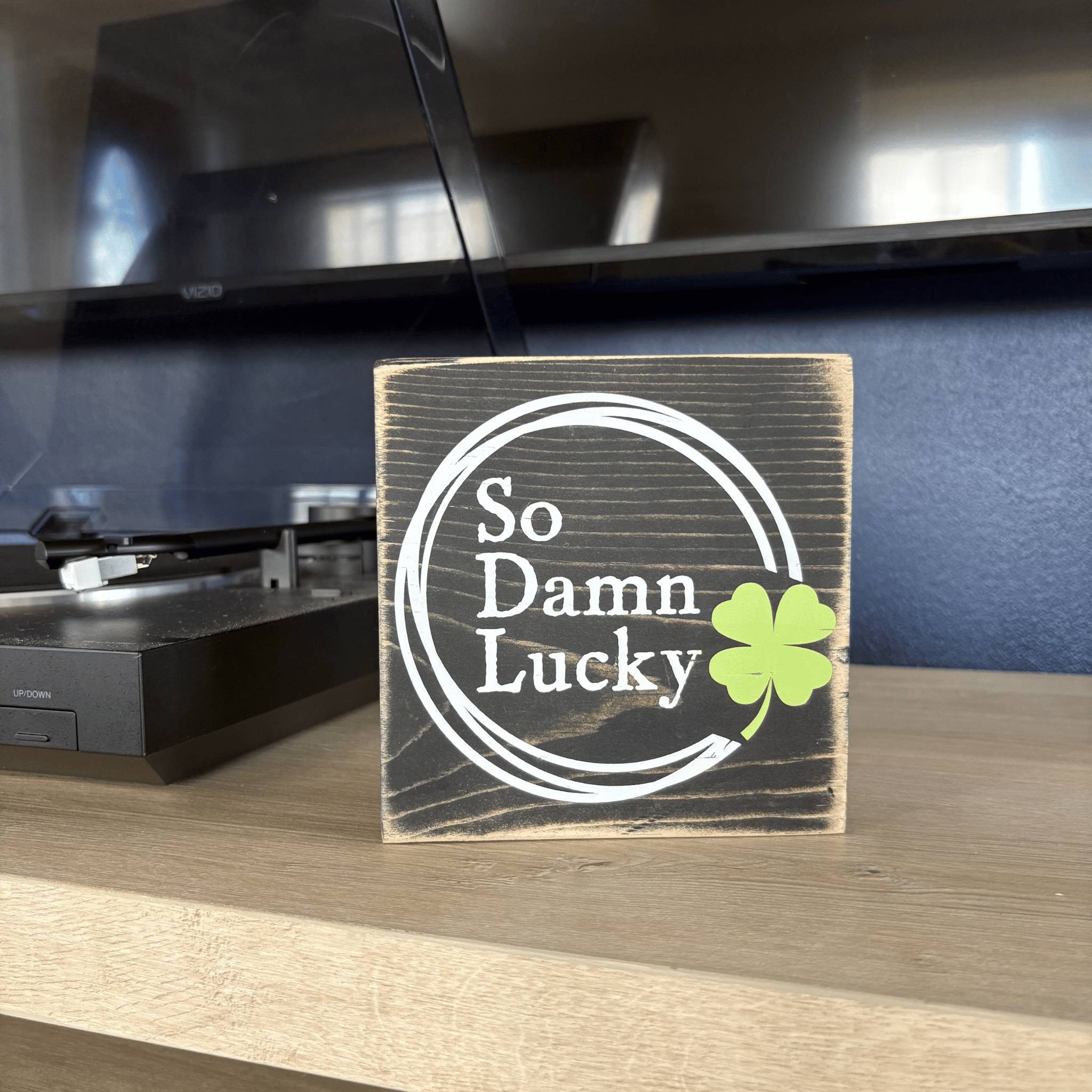 Wooden block with 'So Damn Lucky' and a clover leaf on a table in front of a record player.